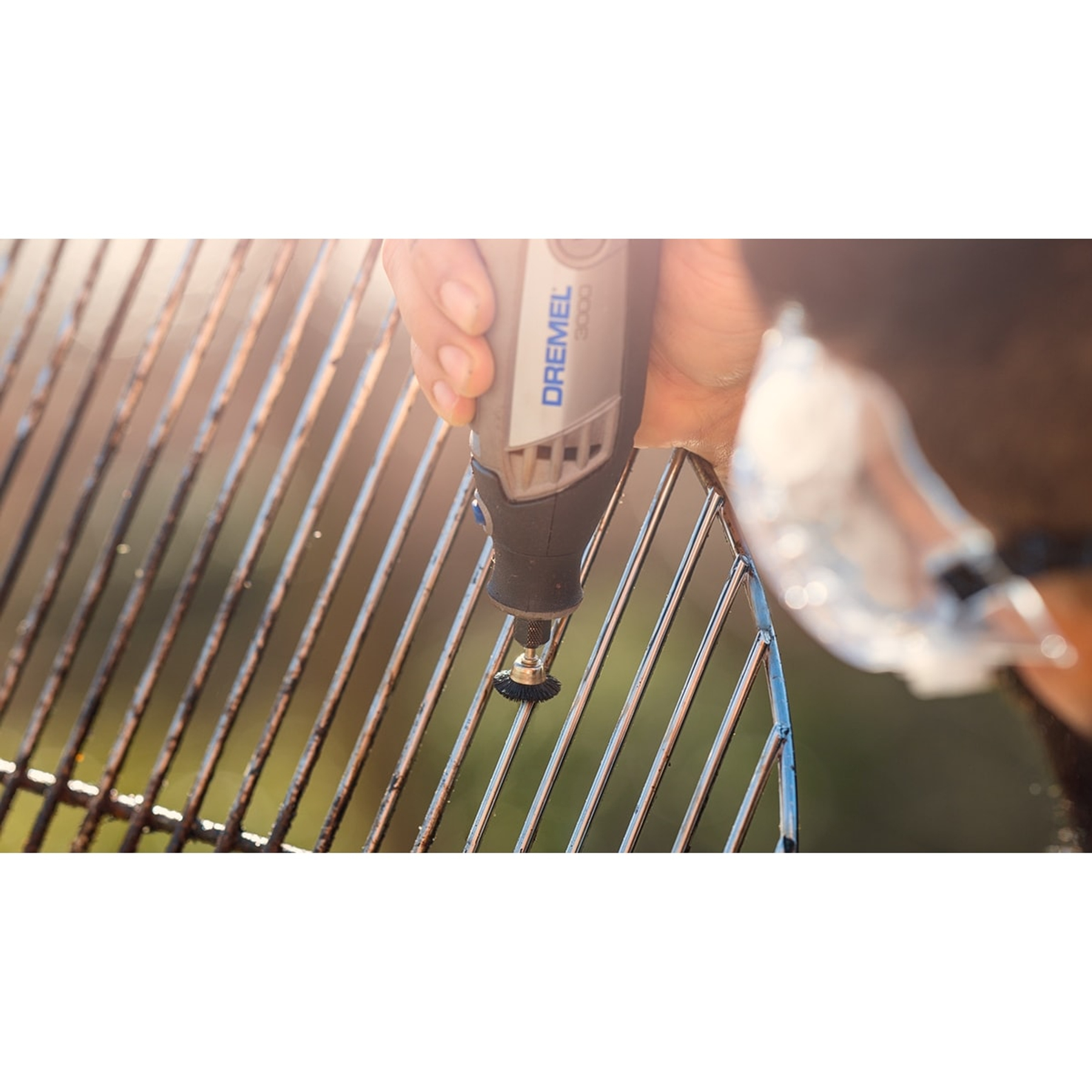 Eine Hand reinigt mit einem Dremel einen rostigen Grill. Das Werkzeug ist grau und schwarz, der Grill ist Stahl, der Hintergrund ist unscharf.
