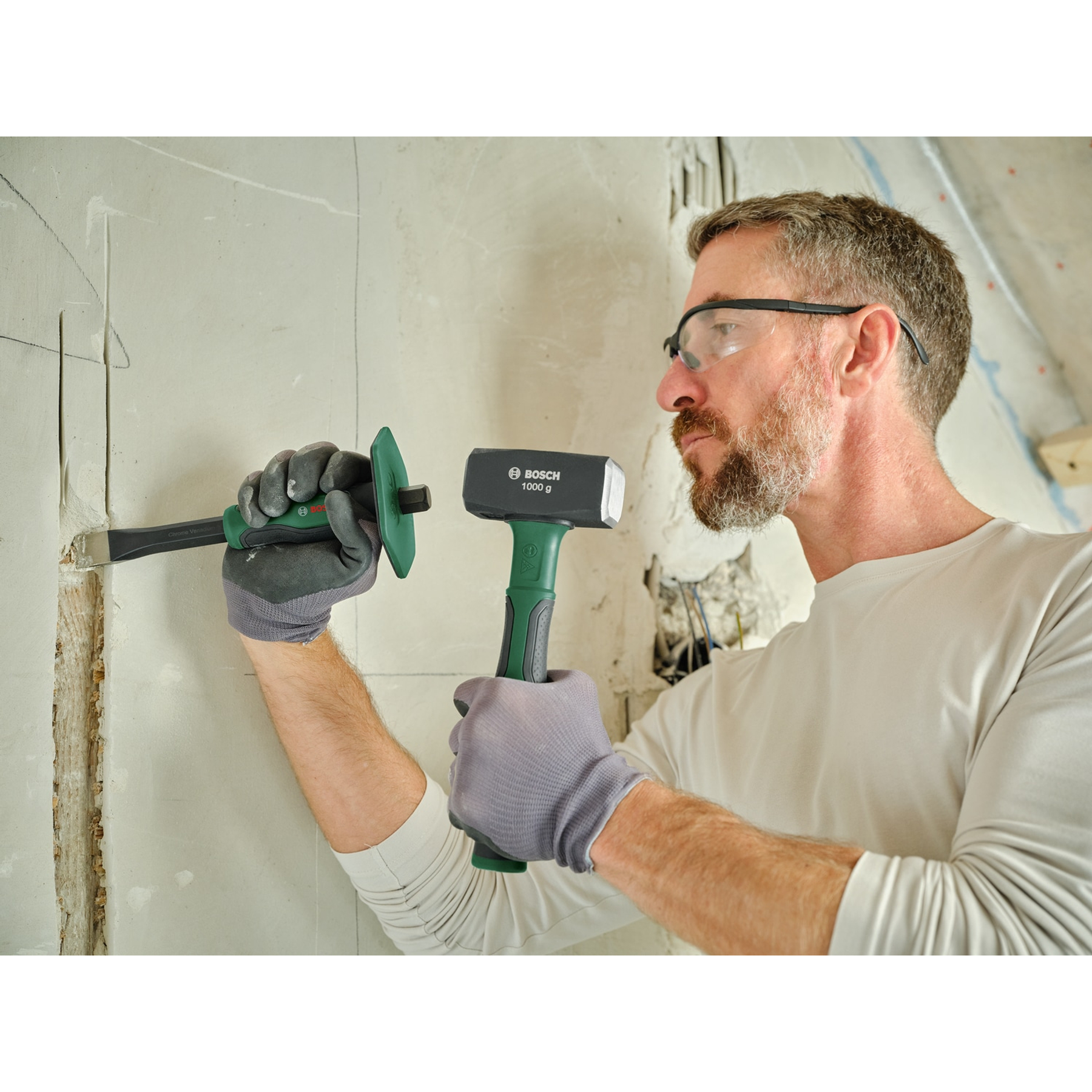 Ein Mann benutzt Meißel und Hammer, um an einer Wand zu arbeiten. Er trägt eine Schutzbrille und Handschuhe, während er arbeitet.