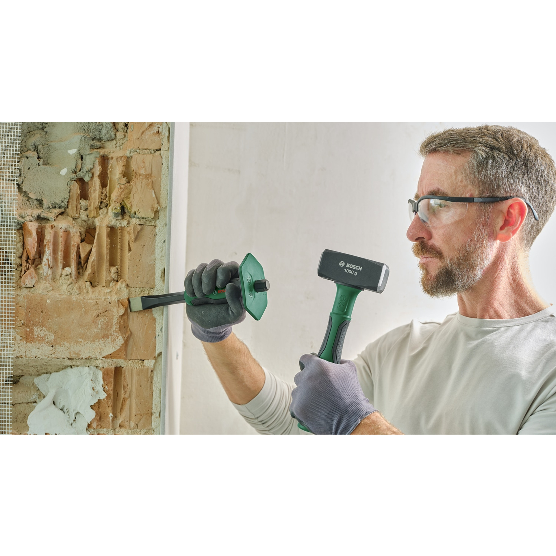 Mann benutzt Meißel und Hammer an einer Ziegelmauer. Er trägt eine Schutzbrille und Handschuhe. Ein Bosch-Hammer ist sichtbar.