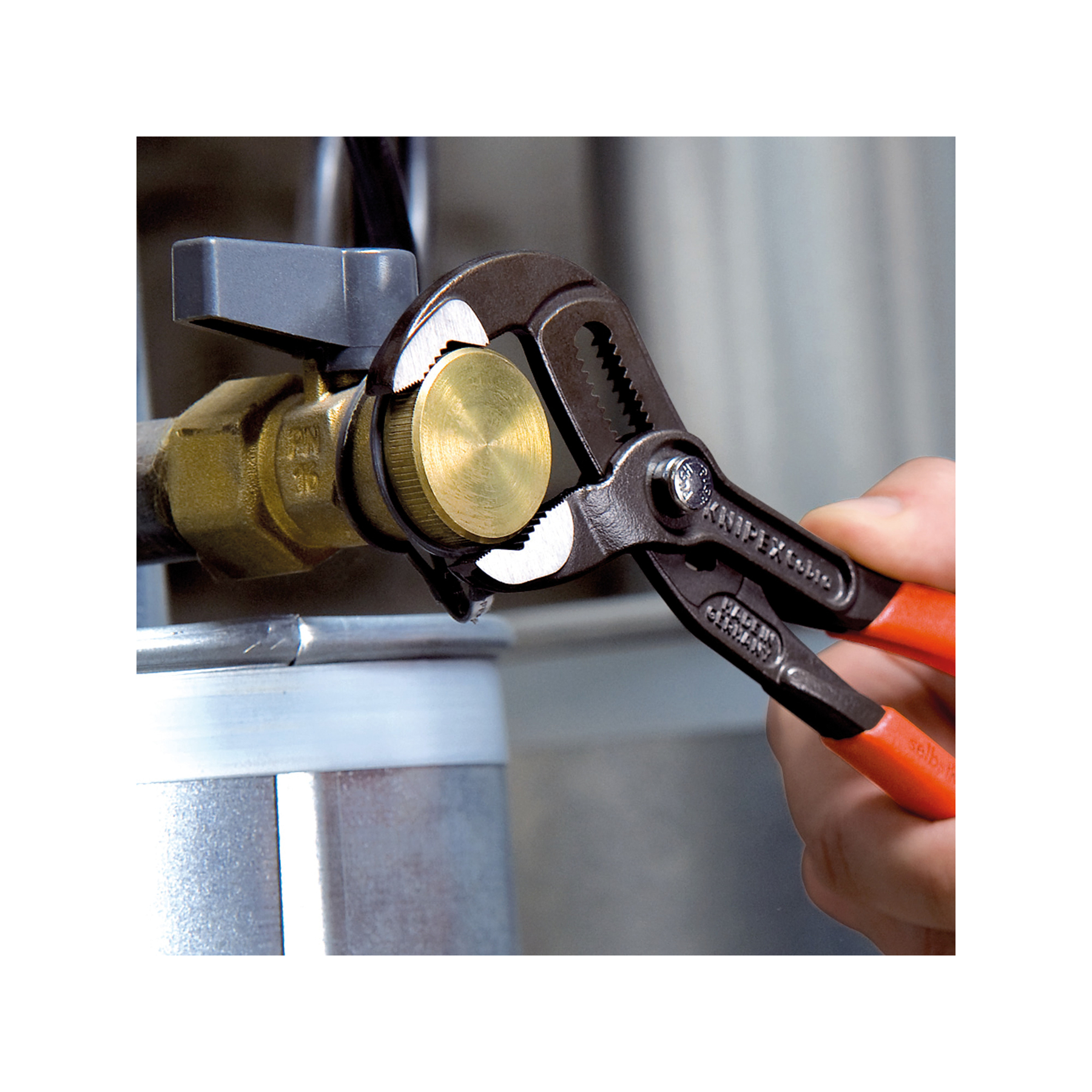 Ręka używa szczypiec do chwytania mosiężnego złącza na rurze. Szczypce są czarne z czerwonymi uchwytami. Szary zawór jest w pobliżu.