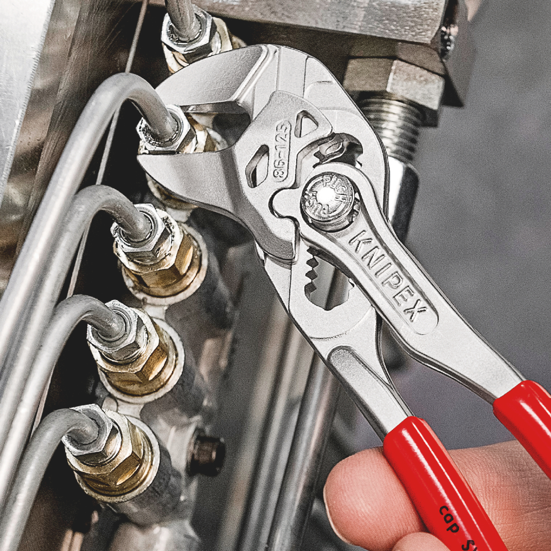 Silver Knipex pliers gripping a metal tube. Red handles are visible. The background includes more metal components.