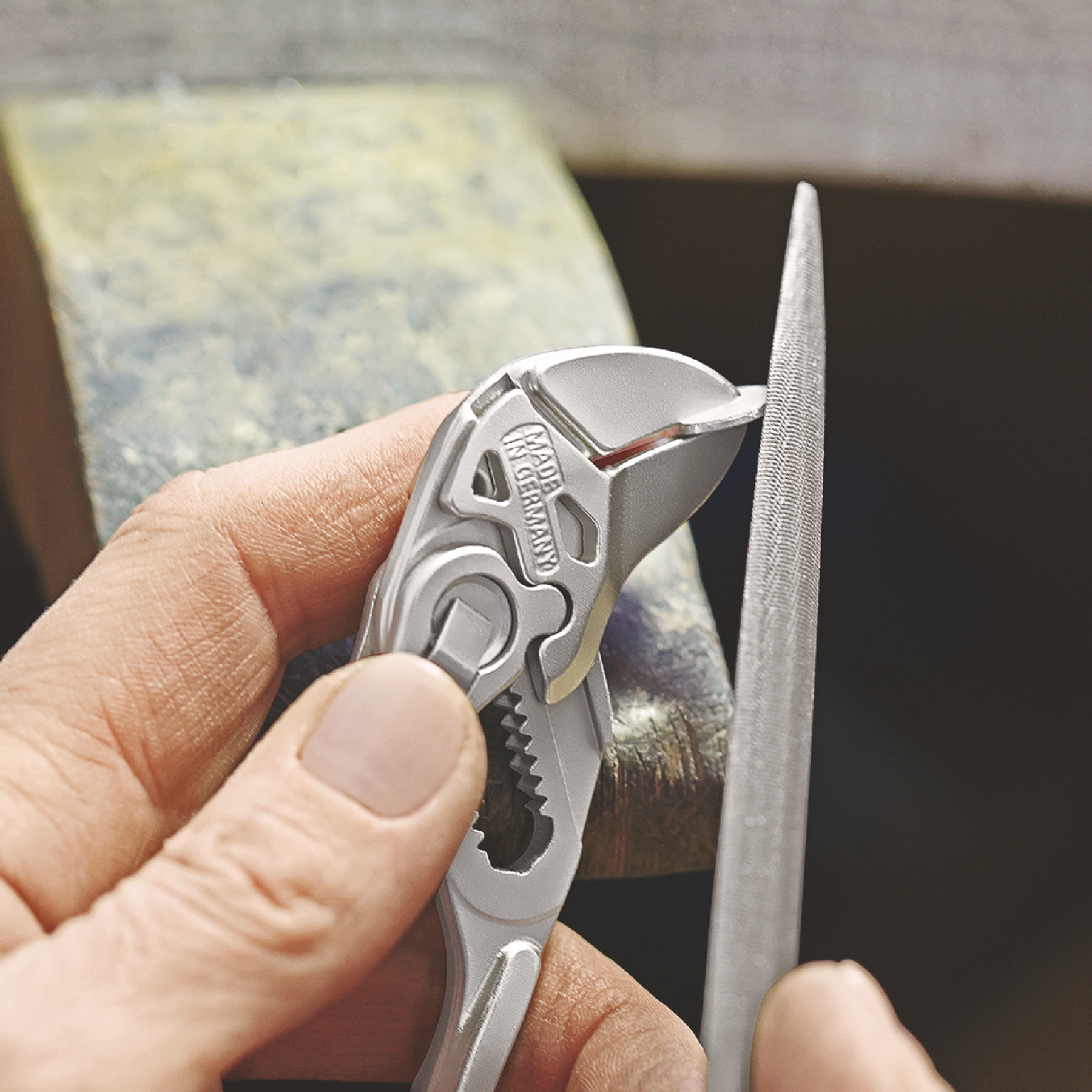 A hand holds pliers against a file. The pliers are close to the file, which is held in place on a work surface.