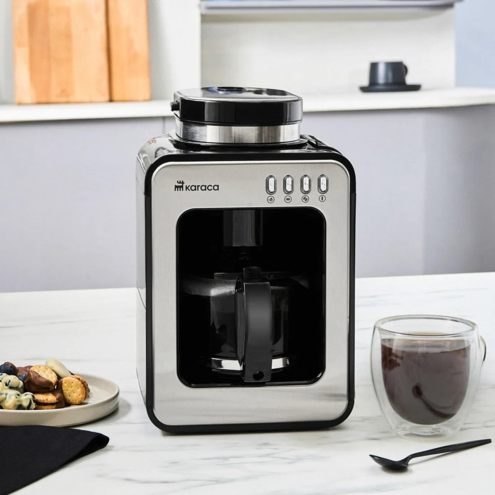 Una macchina per il caffè in acciaio inossidabile con una caraffa di vetro. Accanto c'è una tazza di caffè, con snack su un piatto.