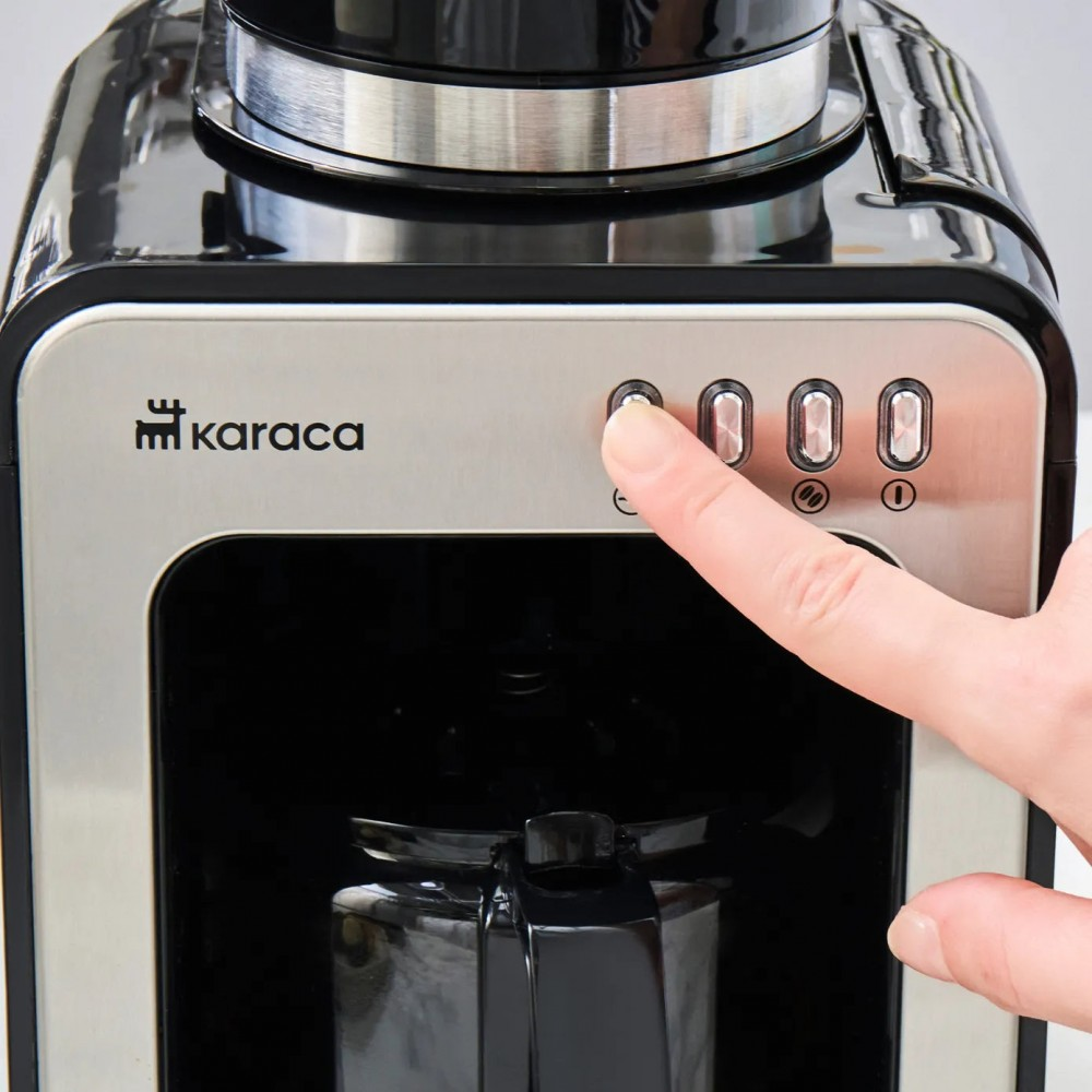 Una mano preme il pulsante di accensione su una macchina da caffè Karaca. La macchina è nera e argento, e lo sfondo è bianco.