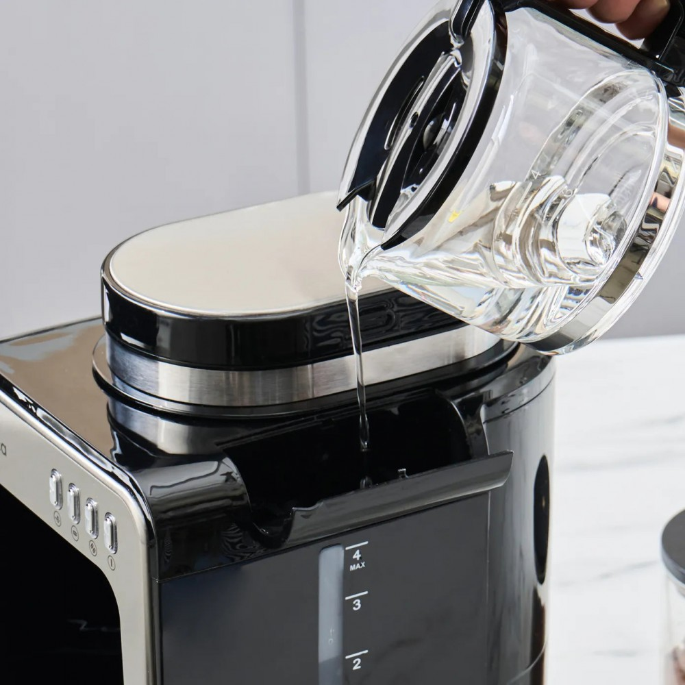 Una persona versa acqua da una brocca trasparente in una macchina da caffè nera e argento, riempiendo il serbatoio dell'acqua.