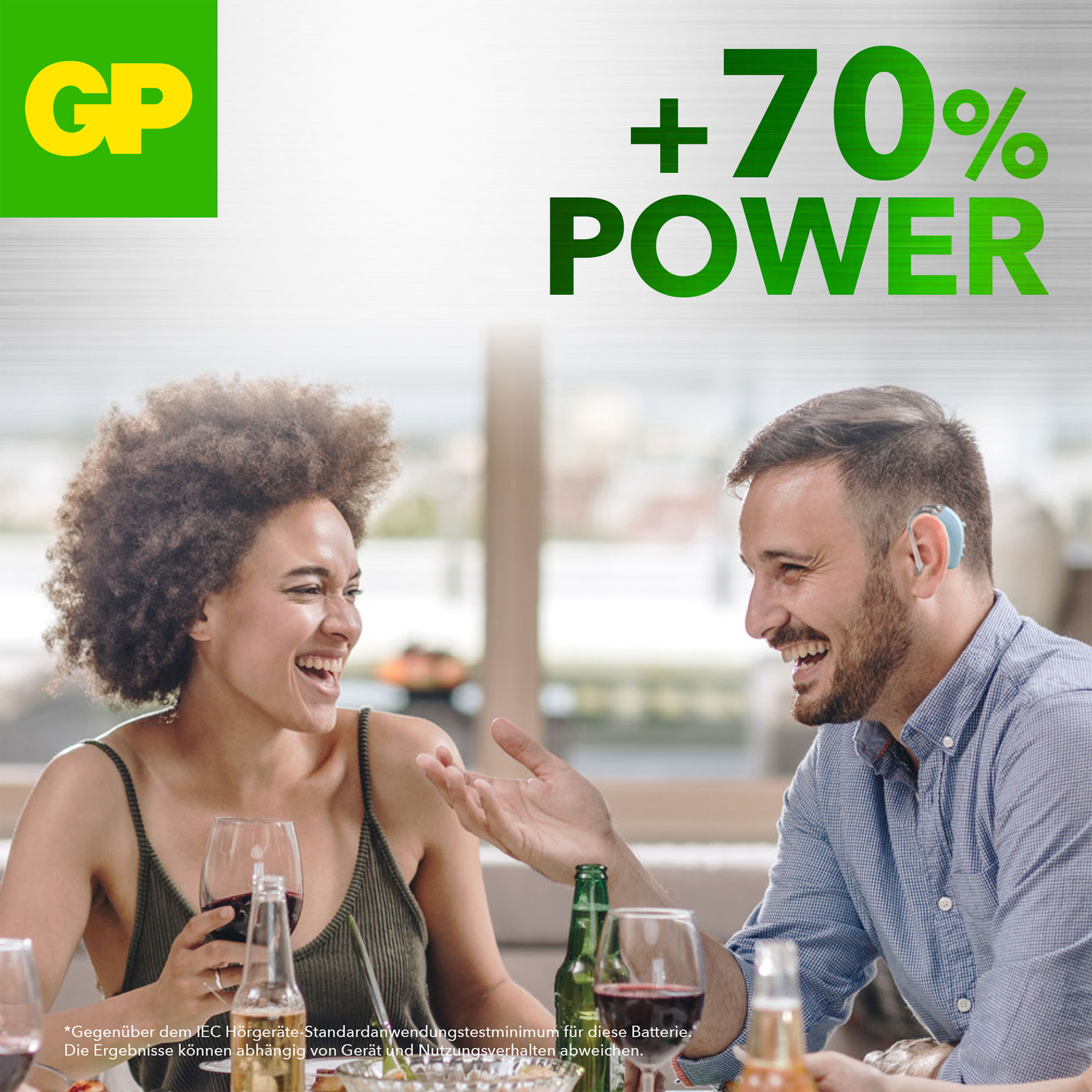 Due persone ridono a tavola. Uno con apparecchio acustico. Logo GP e testo +70% Power sullo sfondo.
