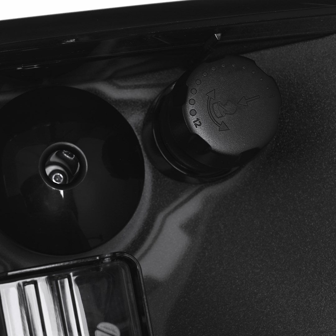 Zbliżenie czarnego elementu maszyny. Zawiera okrągły, przypominający nasadkę przedmiot ze strzałkami, okrągły element i przezroczysty pojemnik.