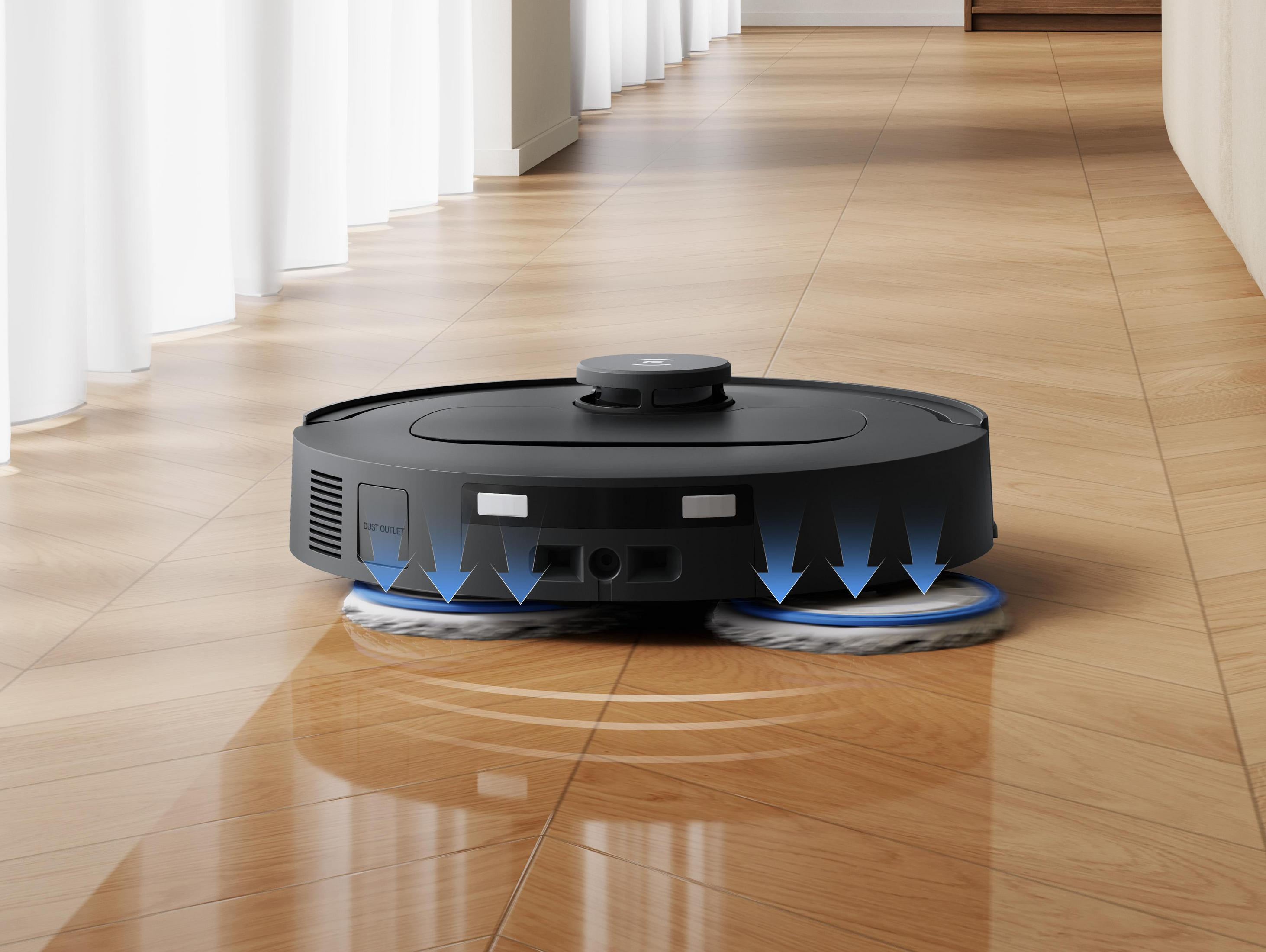 Un aspirapolvere robot nero su pavimento in legno. Ha due mop circolari e tende bianche sullo sfondo.