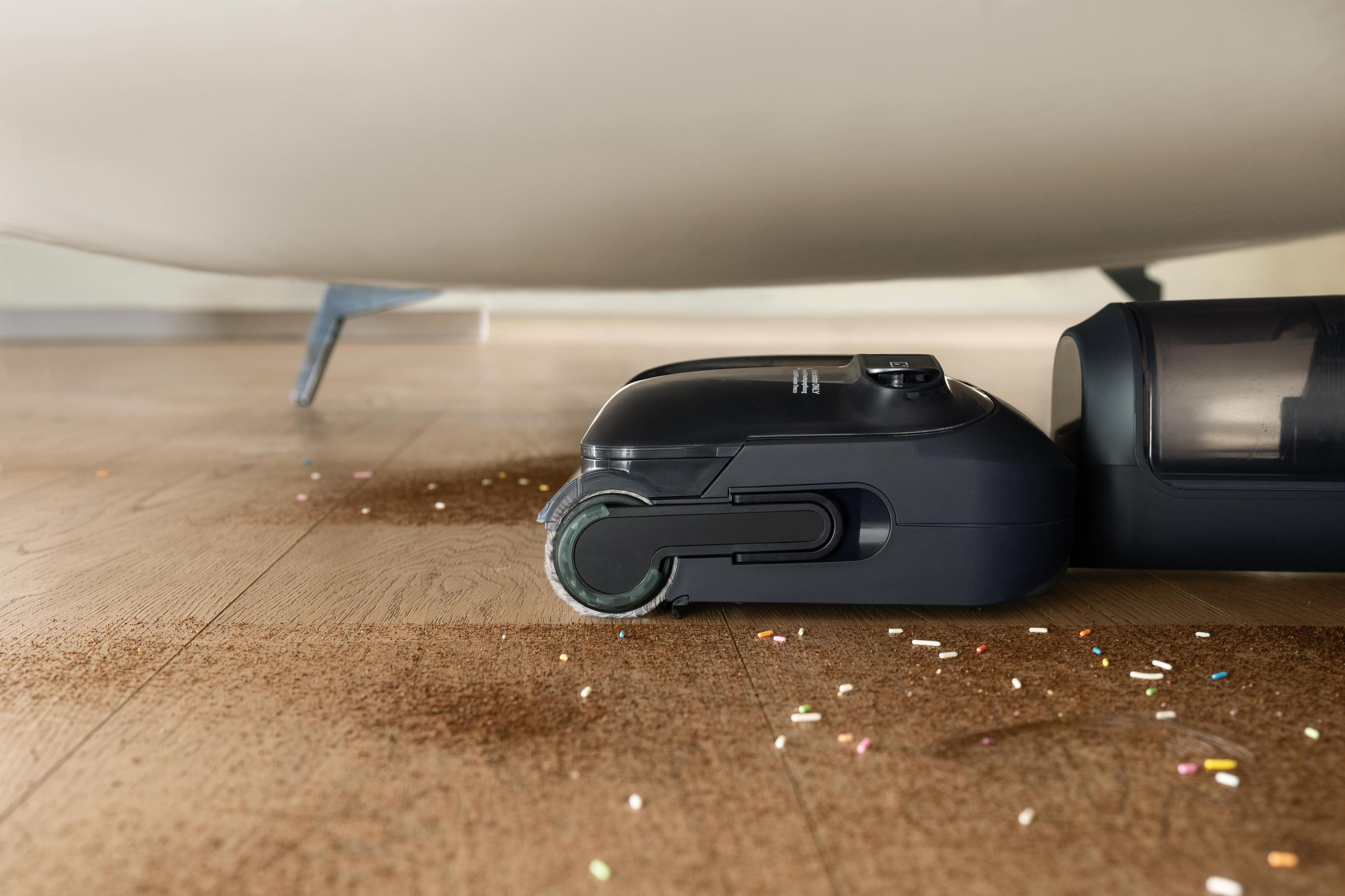 Ciemnoszary odkurzacz robot na drewnianej podłodze pokrytej okruchami i posypką, pod białą powierzchnią.