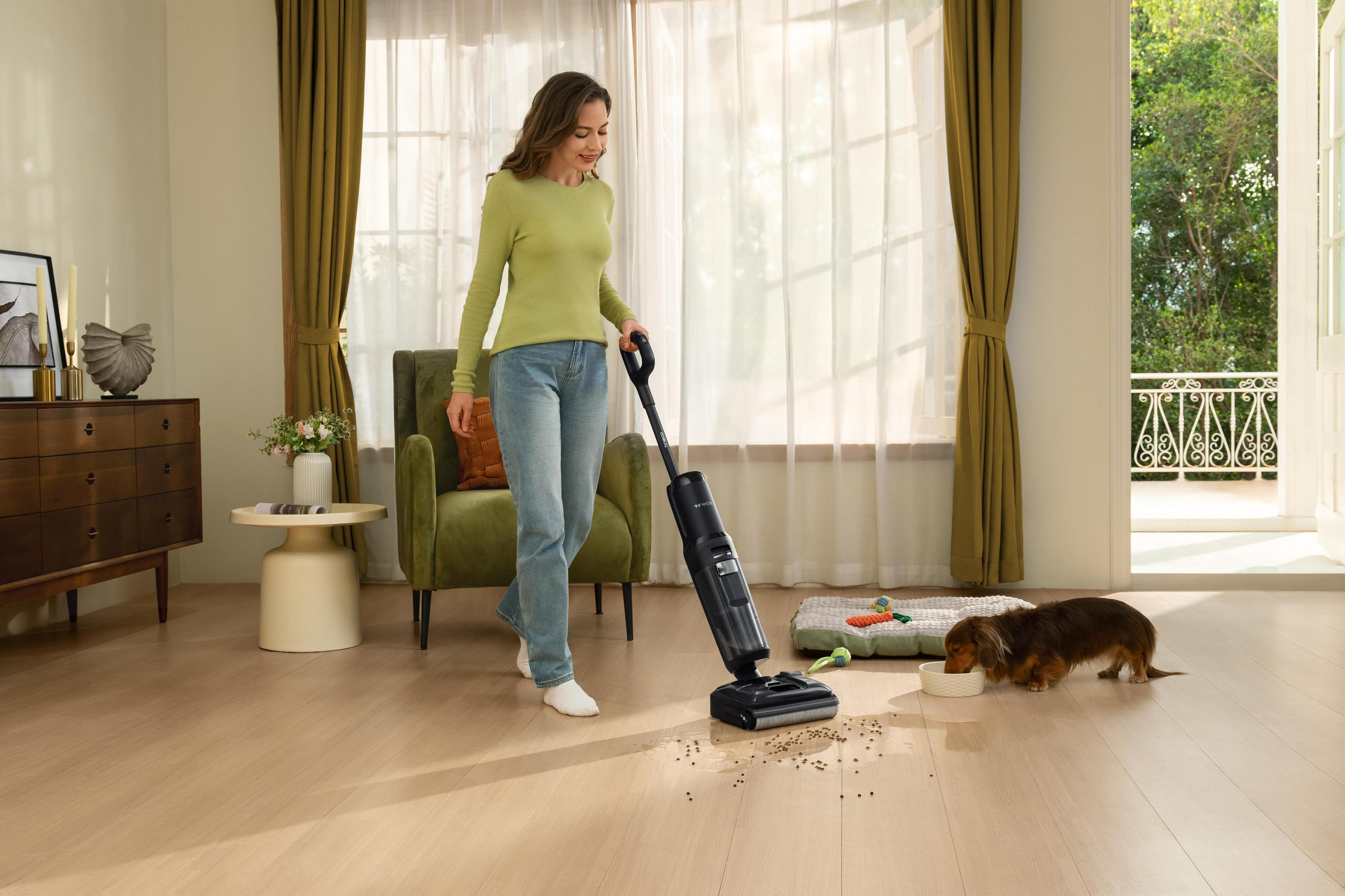 Kobieta odkurza drewnianą podłogę. Pies je z miski. W tle okno i otwarte drzwi.