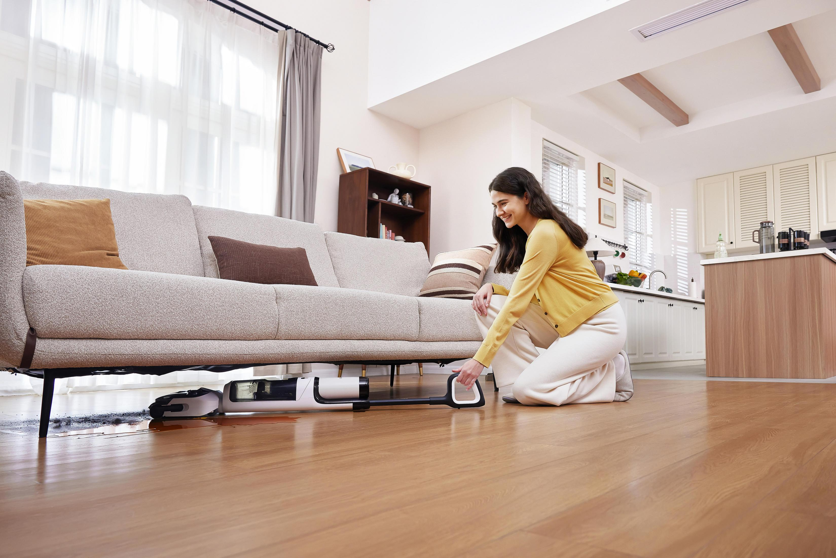 Kobieta klęczy, używając odkurzacza pod kanapą w salonie. Światło wpada przez okno.