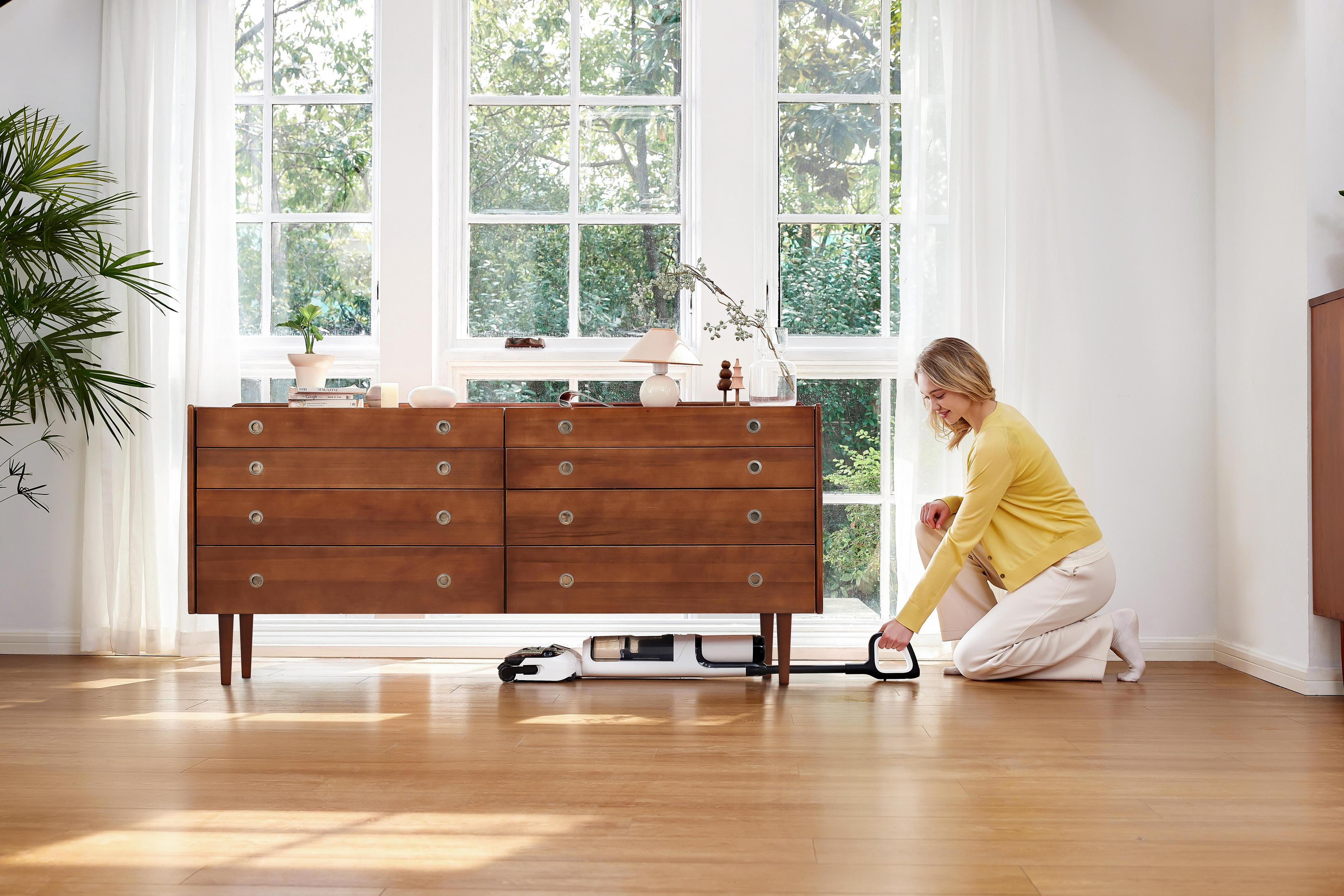 Kobieta klęczy, obsługując myjkę do podłóg. Drewniana komoda obok okna. Rośliny i światło wpadające.