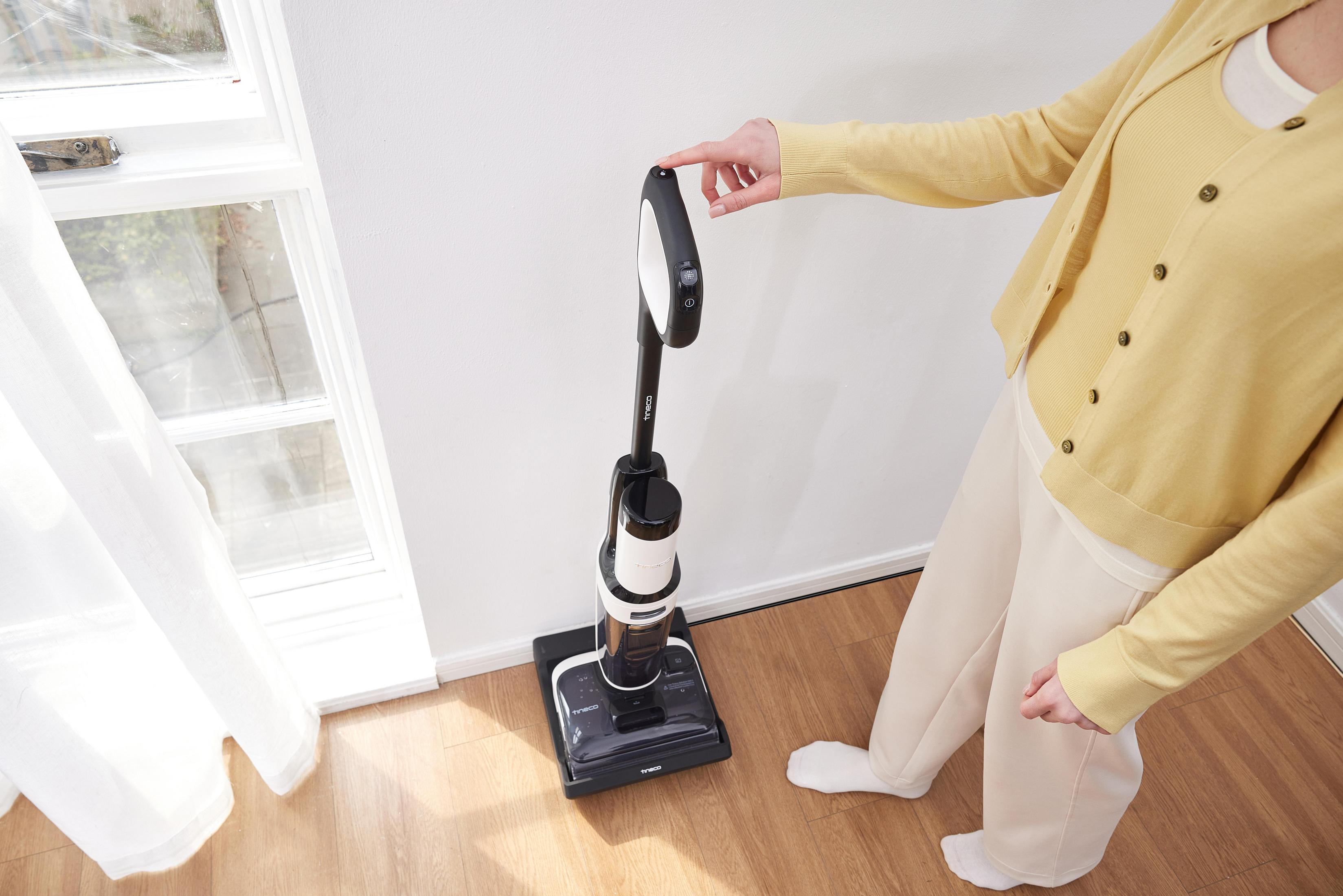 Kobieta w żółtym kardiganie obsługuje czarno-biały odkurzacz w pokoju z oknem.