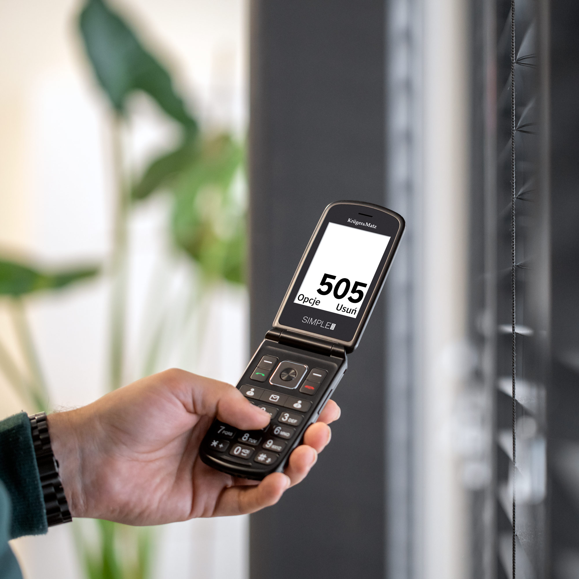 Dłoń trzyma otwarty czarny telefon komórkowy z klapką z numerem 505. Tło obejmuje okno i roślinę.
