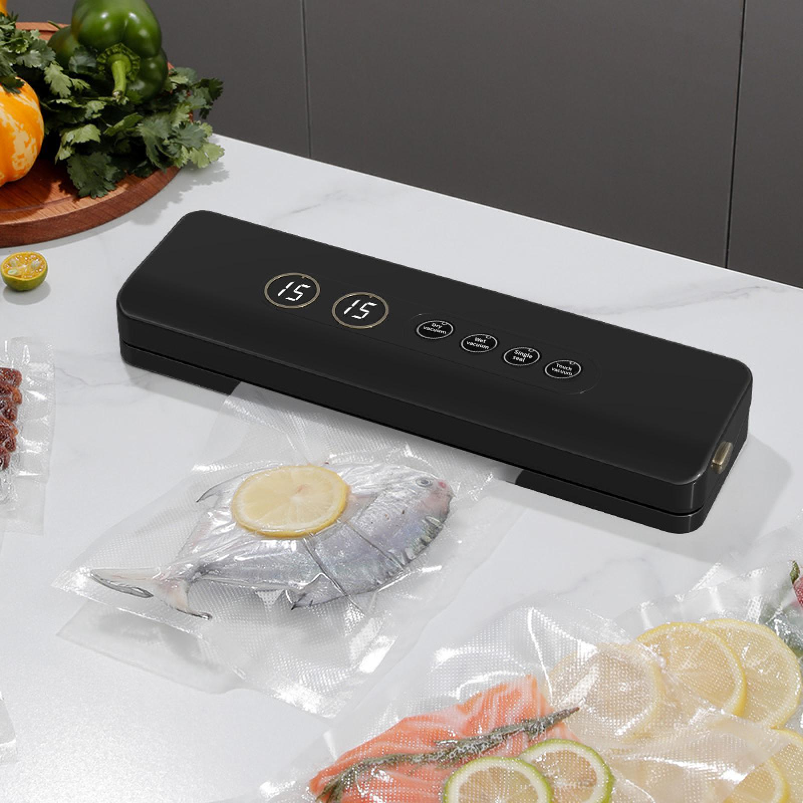 Eine schwarze Vakuumier maschine auf einer weißen Oberfläche mit versiegeltem Fisch, Zitronenscheiben und Gemüse im Hintergrund.