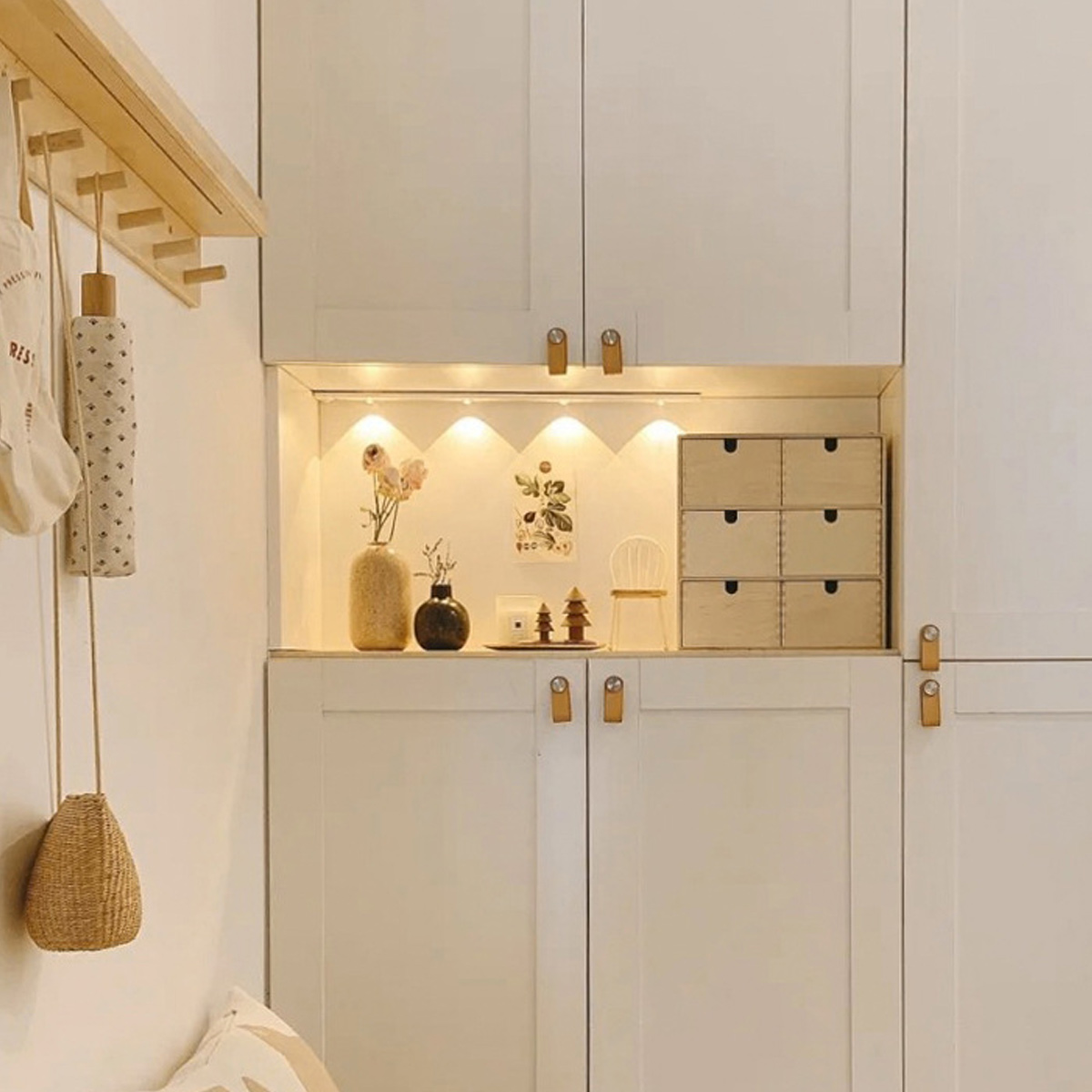 Ein weißer Schrank mit Schubladen, Vasen und Dekorationen. Ein Holzregal hält eine Tasche und einen Regenschirm. Ein kleiner Stuhl und Holzkisten sind sichtbar.