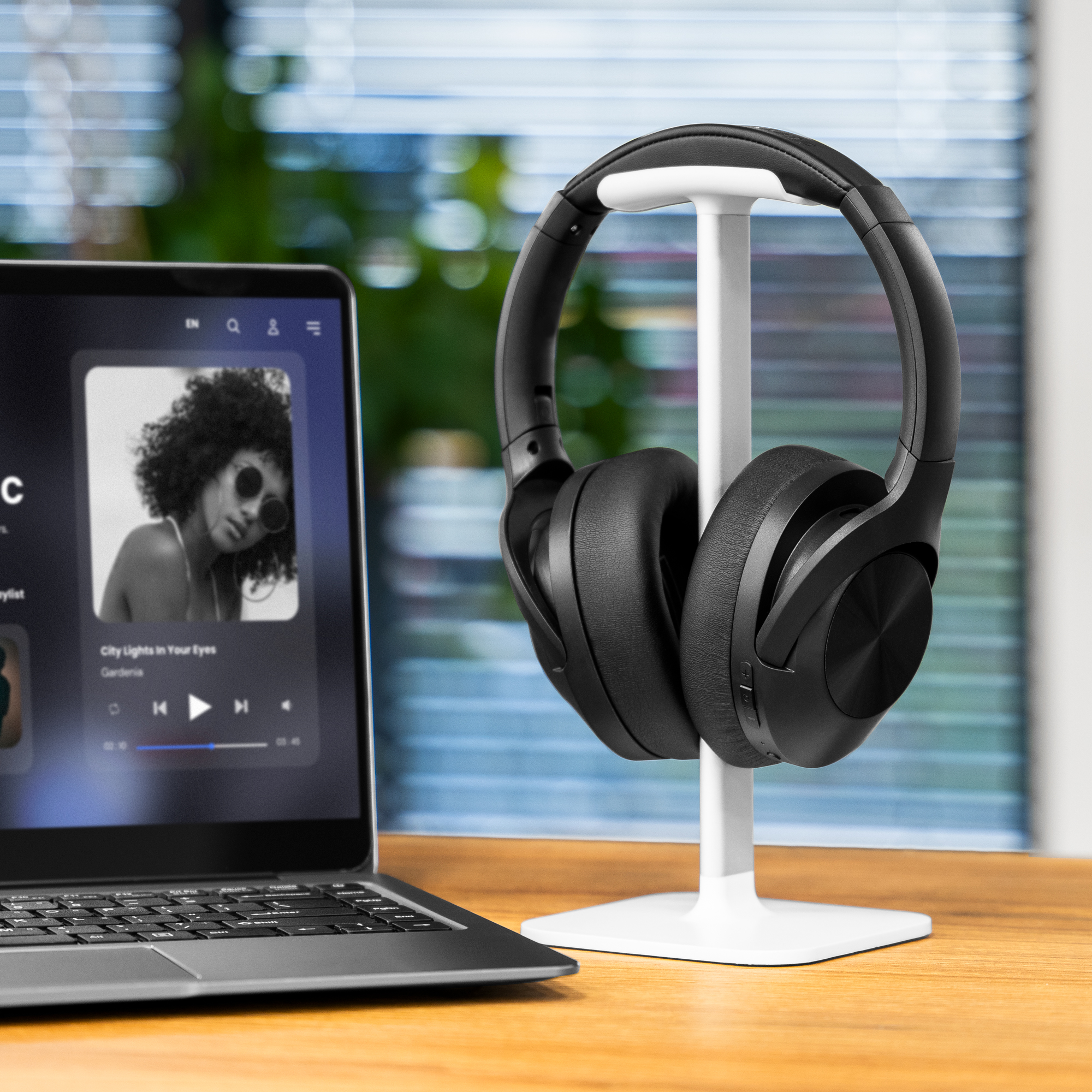 Czarne słuchawki na białym stojaku, laptop odtwarzający muzykę ze zdjęciem kobiety, drewniane biurko, rozmyte tło.