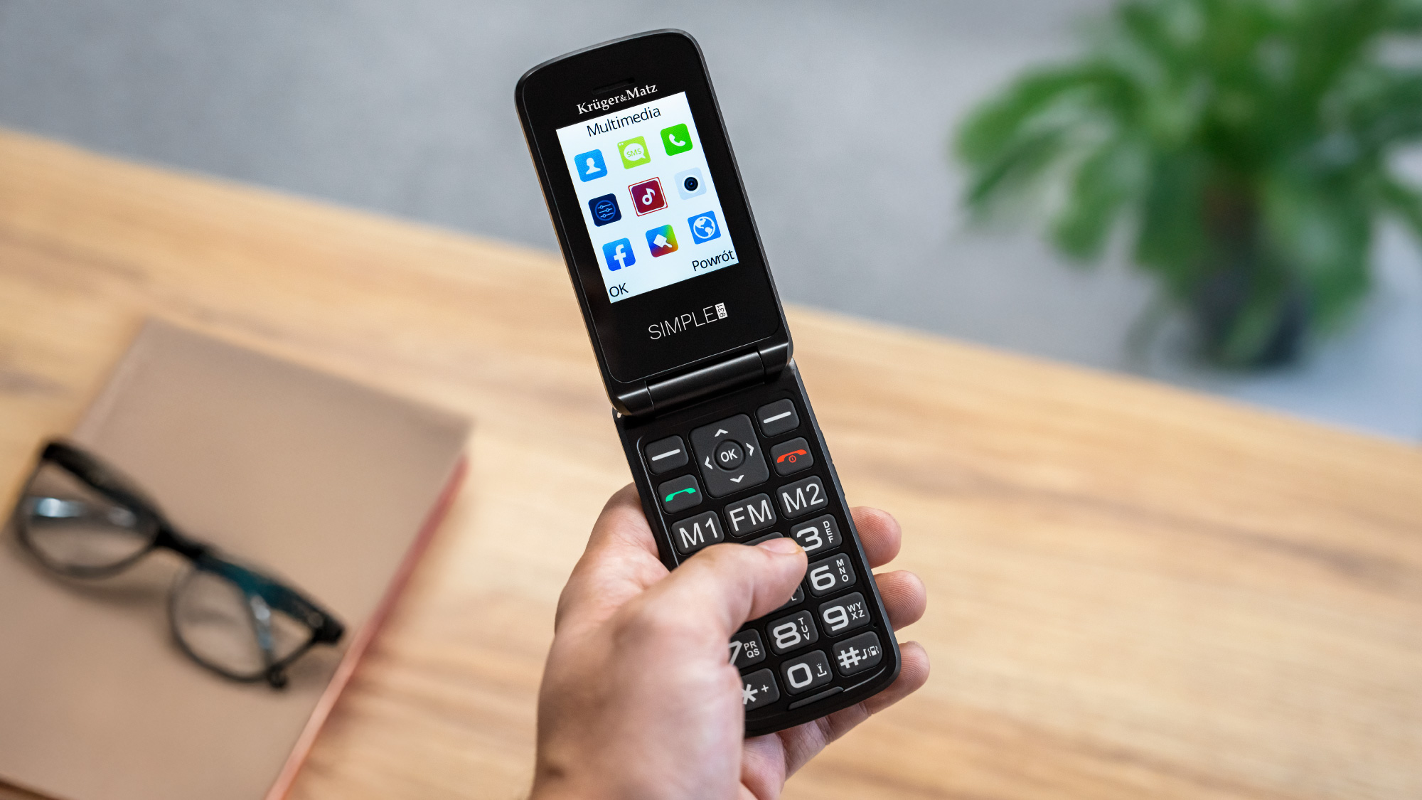 Ręka trzyma czarny telefon z klapką z otwartym ekranem, pokazującym ikony aplikacji. Książka i okulary leżą na drewnianym stole.