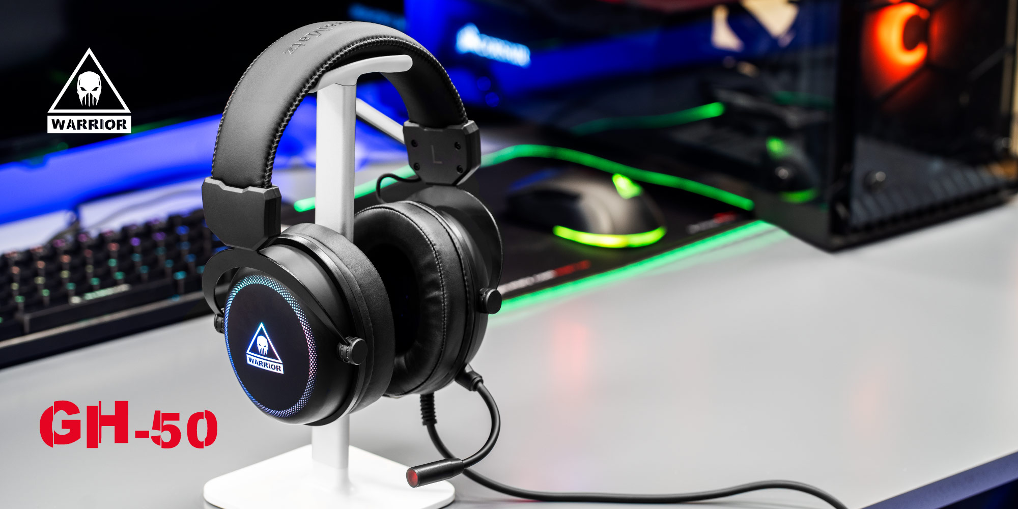 Czarne słuchawki dla graczy na białym stojaku. Klawiatura, mysz i komputer w tle. Model słuchawek to GH-50.