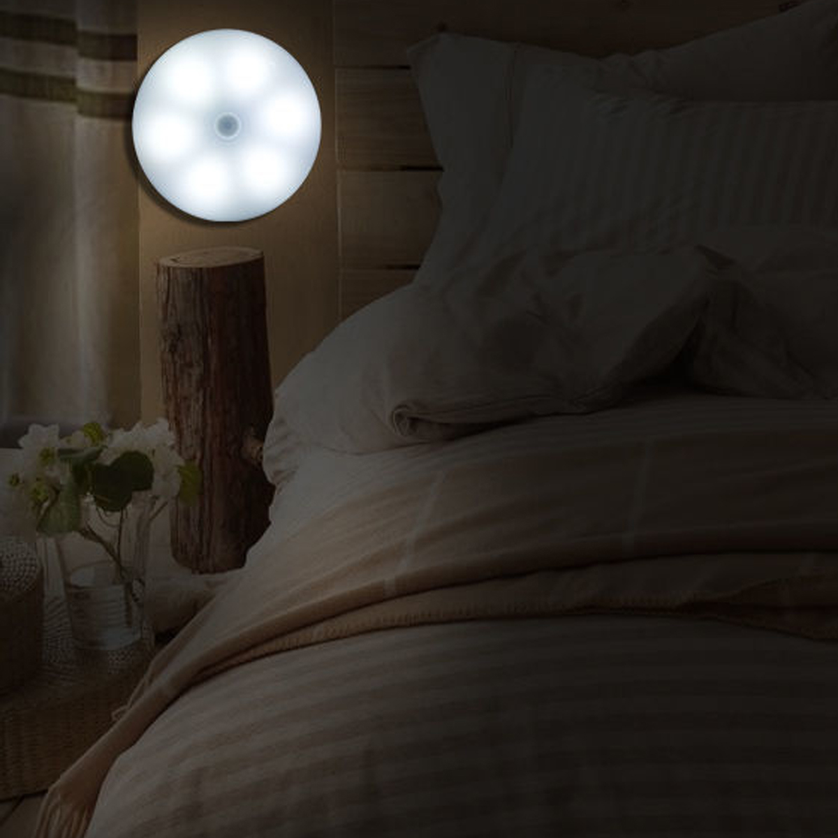 Eine beleuchtete runde LED-Lampe an einem hölzernen Bettrahmen. Ein Bett mit weißer Bettwäsche. Ein Baumstamm und Blumen sind links.
