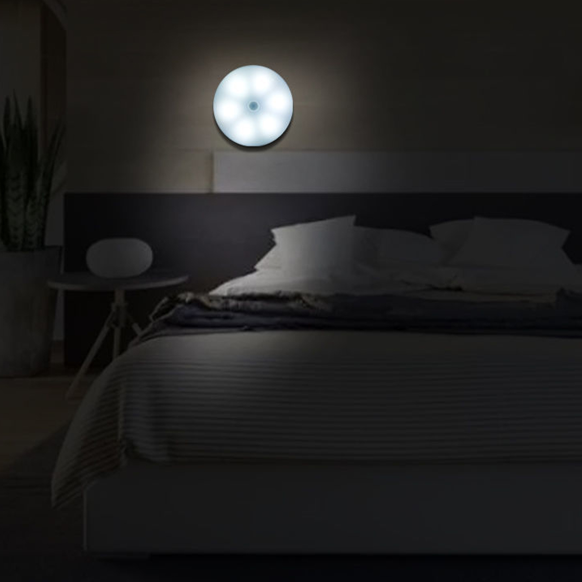 Eine beleuchtete, runde Lampe an einer dunklen Wand über einem Bett. Das Bett ist mit weißen Kissen und einer grauen Decke gemacht. Daneben steht ein kleiner Tisch.