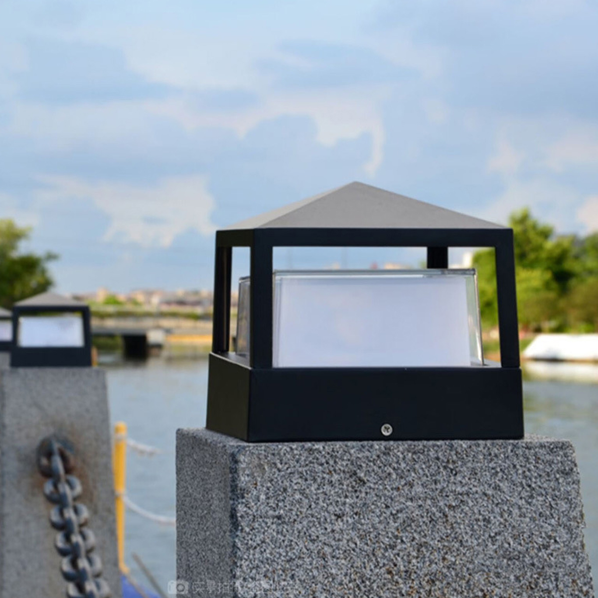 Schwarze Lampe auf Granitsäule. Eine zweite Lampe ist im Hintergrund unscharf. Ein Fluss und Himmel mit Wolken sind sichtbar.