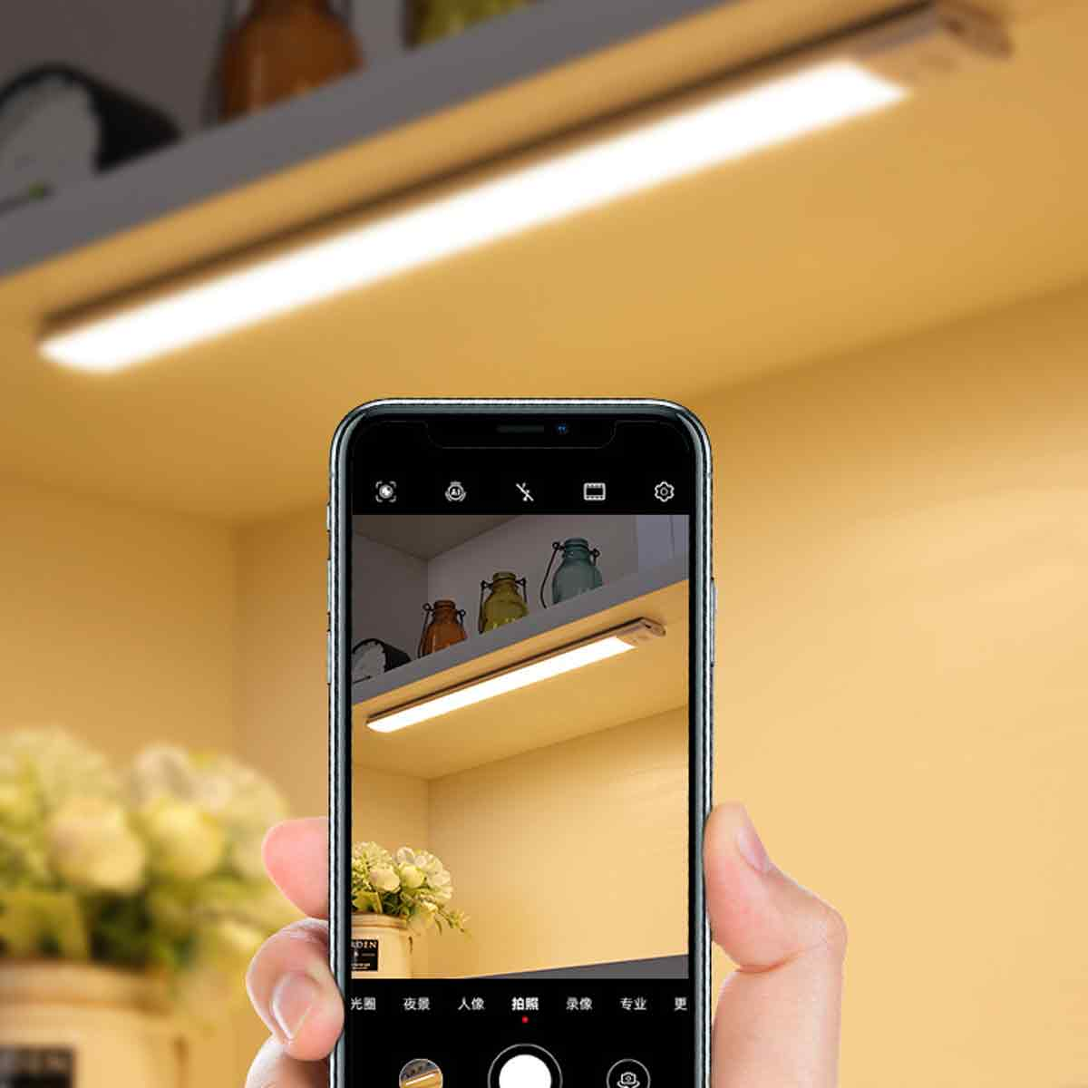 Eine Hand hält ein Telefon, das eine Leuchte fotografiert. Im Hintergrund sind ein Regal mit Gläsern und Blumen zu sehen.