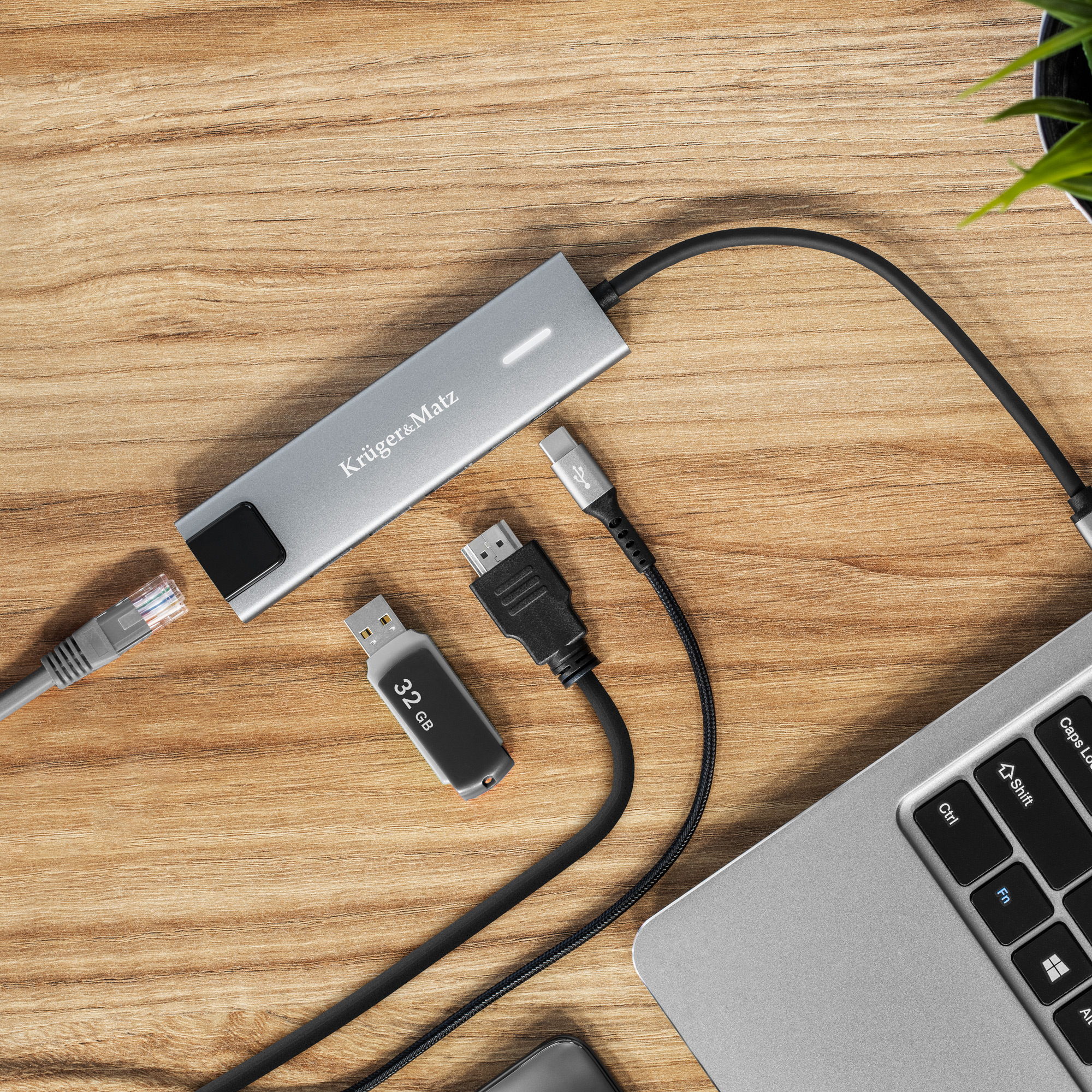 Drewniane biurko ze srebrnym hubem, pendrivem i laptopem. Kable łączą te urządzenia. Mała roślina w tle.