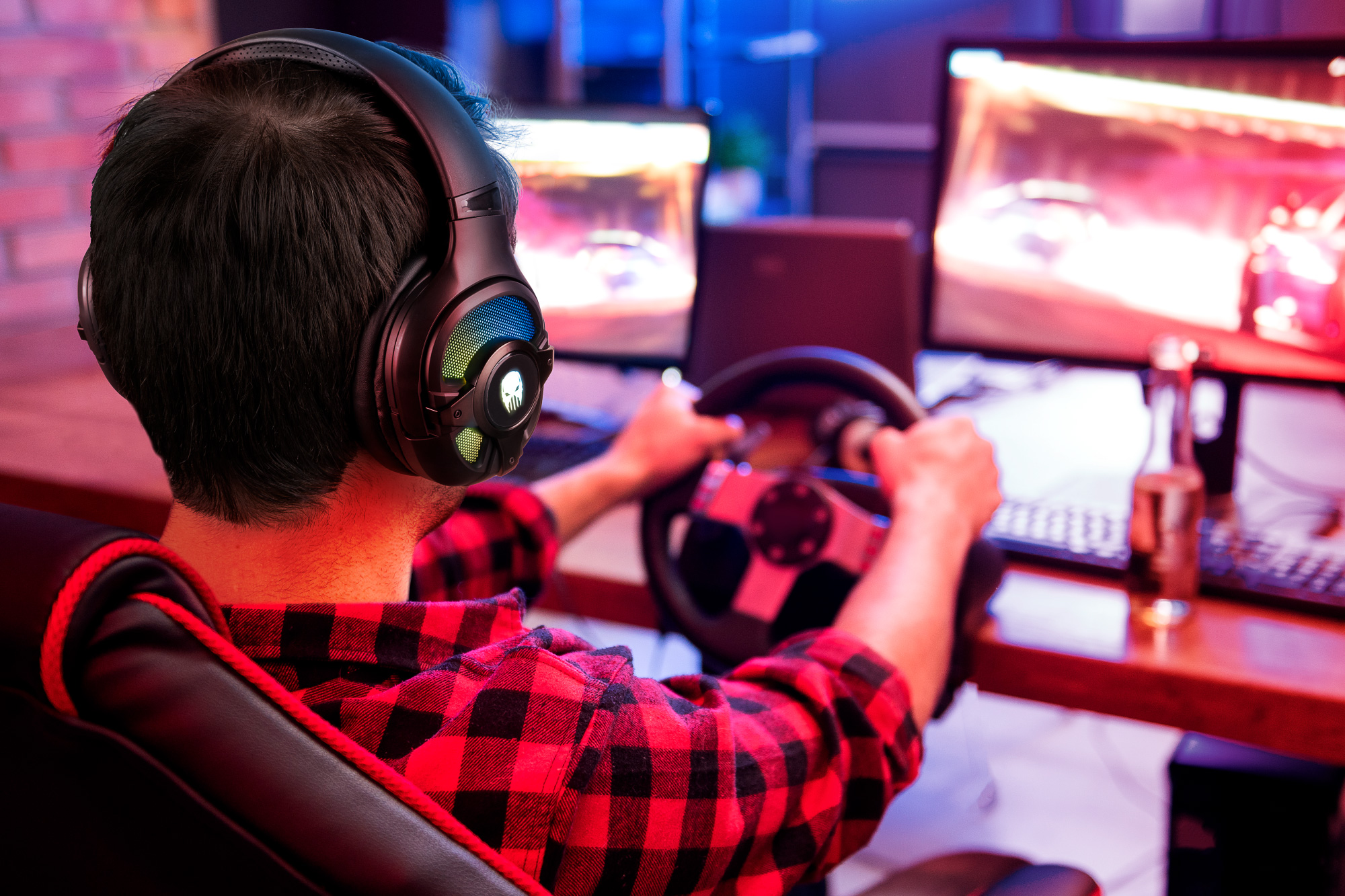Osoba w słuchawkach gra w grę wyścigową za pomocą kierownicy. Ma na sobie czerwoną koszulę w kratę. Widoczne są monitory.