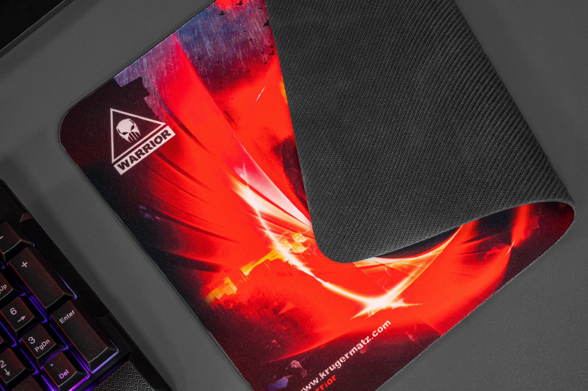 Podkładka pod mysz dla graczy z logo czaszki i napisem 'Warrior'. Jest czerwono-czarna, obok klawiatury. Podkładka jest częściowo zwinięta.