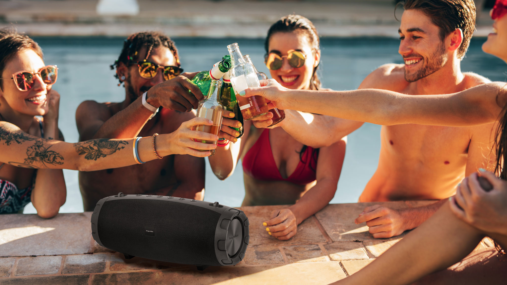 Ludzie wznoszą toast butelkami przy basenie. Czarny głośnik leży na brzegu. Widoczne okulary przeciwsłoneczne, tatuaże i uśmiechy.