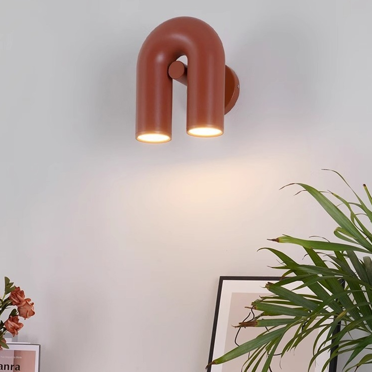 Eine rostfarbene Wandleuchte mit zwei nach unten gerichteten Lichtern ist an einer weißen Wand angebracht. Pflanzen und ein gerahmtes Bild befinden sich im Vordergrund.