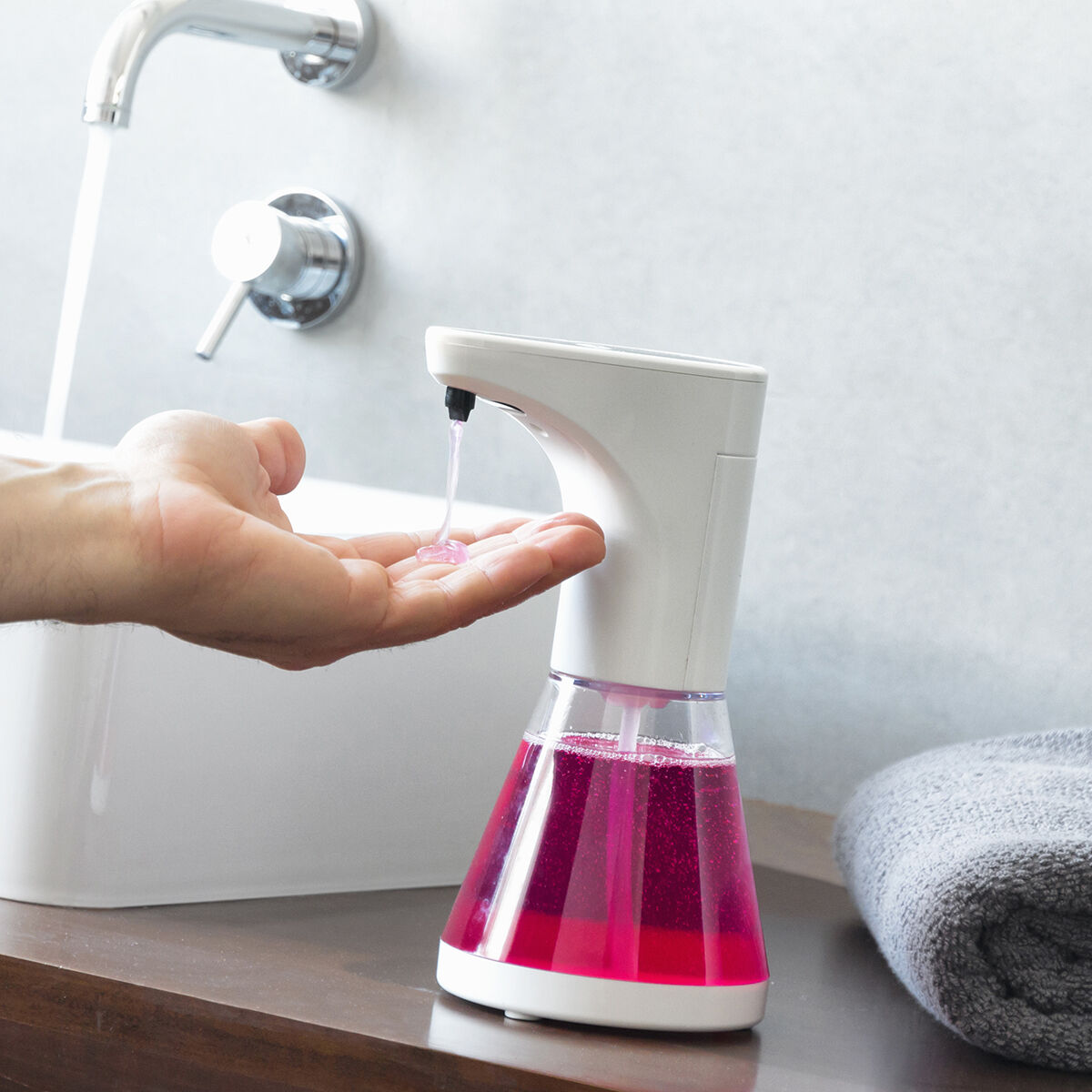 Eine Hand empfängt rosa Seife aus einem automatischen Spender. Eine Badezimmerszene mit Waschbecken, Wasserhahn und einem grauen Handtuch.
