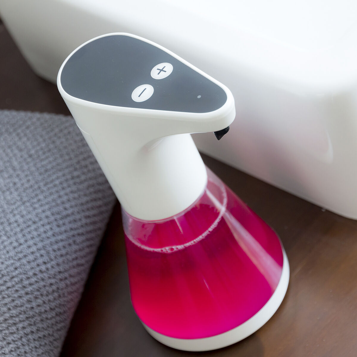 Ein weiß-schwarzer automatischer Seifenspender mit rosa Flüssigkeit. Im Hintergrund ein graues Handtuch und eine weiße Oberfläche.