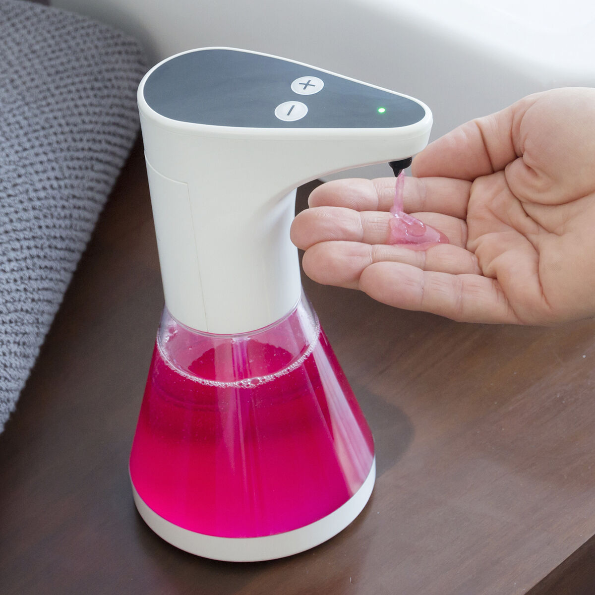 Ein weiß-schwarzer automatischer Seifenspender gibt rosa Seife in eine Hand ab. Der Spender steht auf einer braunen Oberfläche, Badezimmer im Hintergrund.