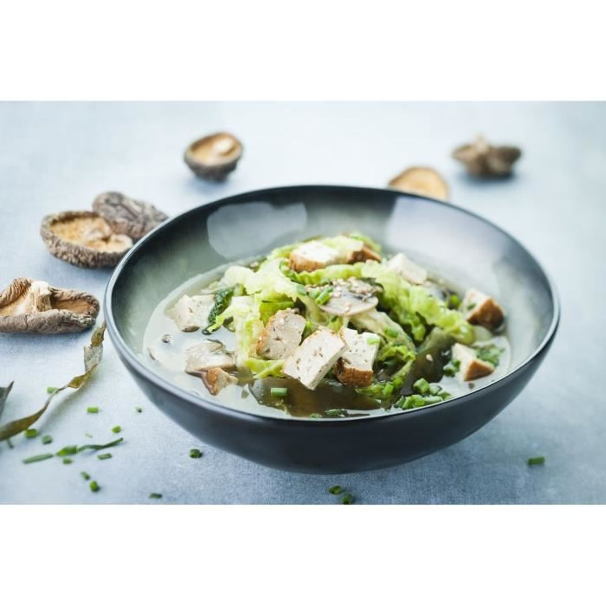 Eine Schüssel Miso-Suppe mit Tofu, Pilzen und grünem Gemüse. Getrocknete Pilze sind auf einer hellblauen Oberfläche verstreut.