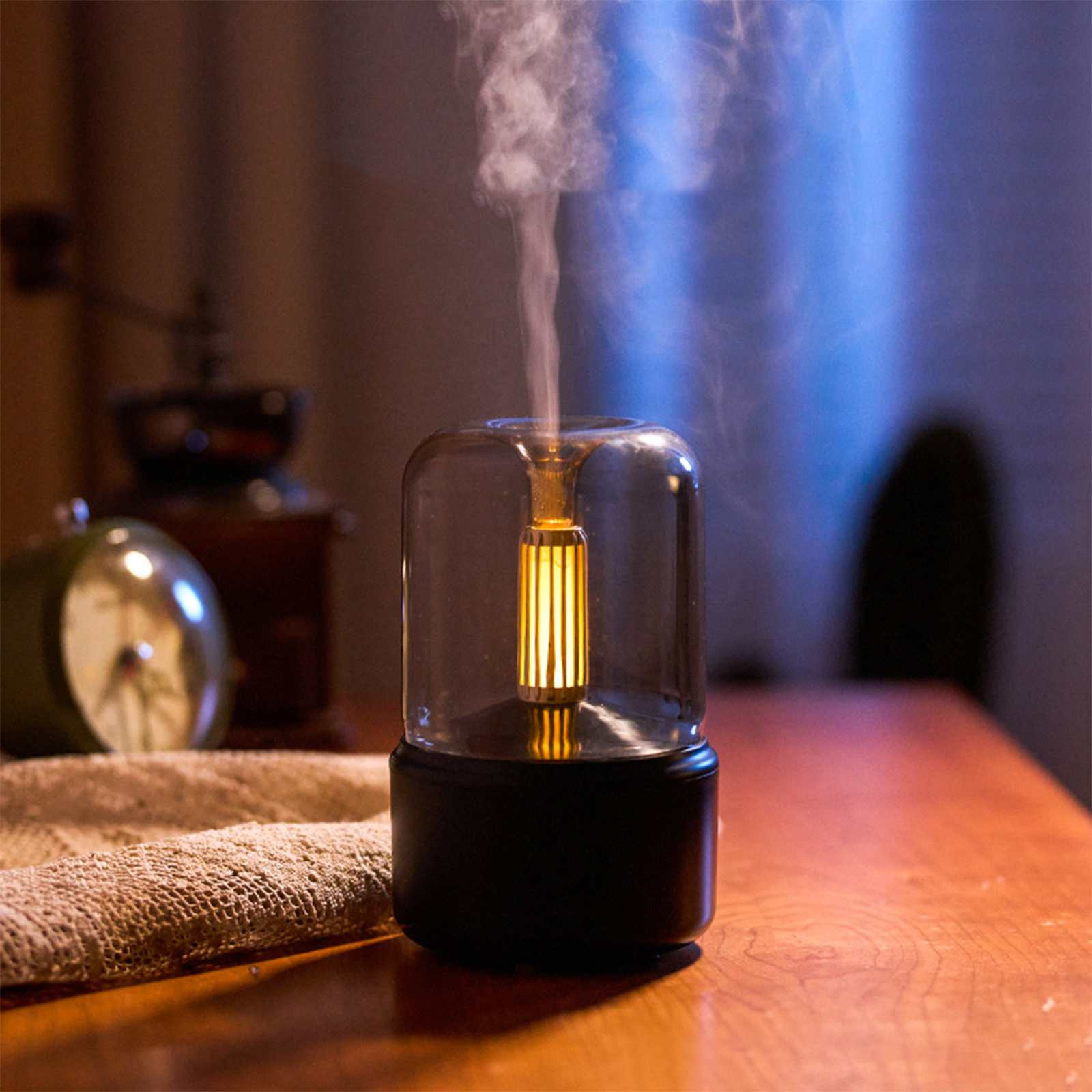 Ein beleuchteter Luftbefeuchter mit Glasabdeckung steht auf einem Holztisch. Dampf steigt daraus auf. Eine Uhr und eine Mühle sind im Hintergrund sichtbar.
