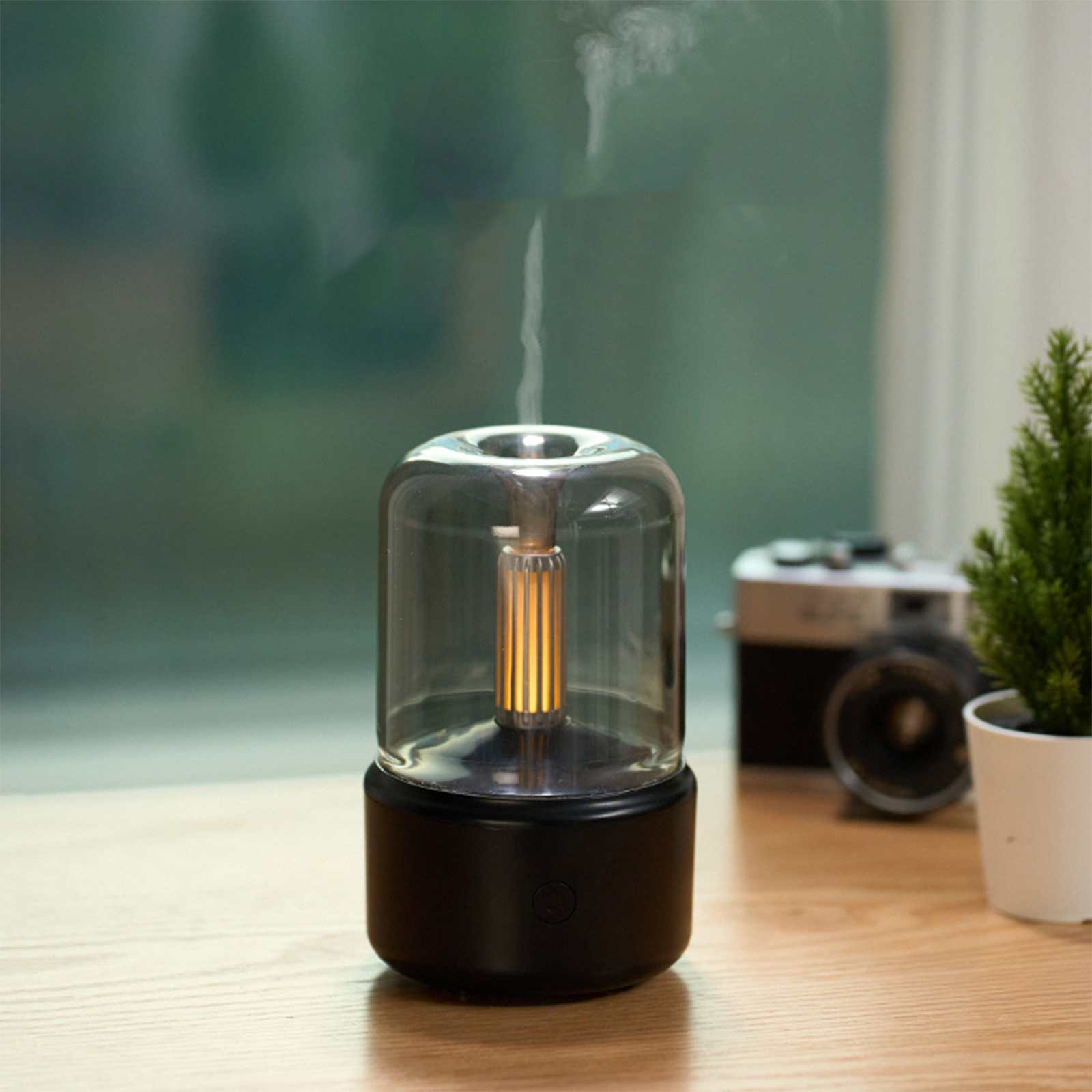 Auf einer Holzoberfläche befindet sich ein schwarzer Glasdiffusor. Ein kleiner Baum und eine Kamera befinden sich im Hintergrund. Rauch steigt aus dem Diffusor auf.