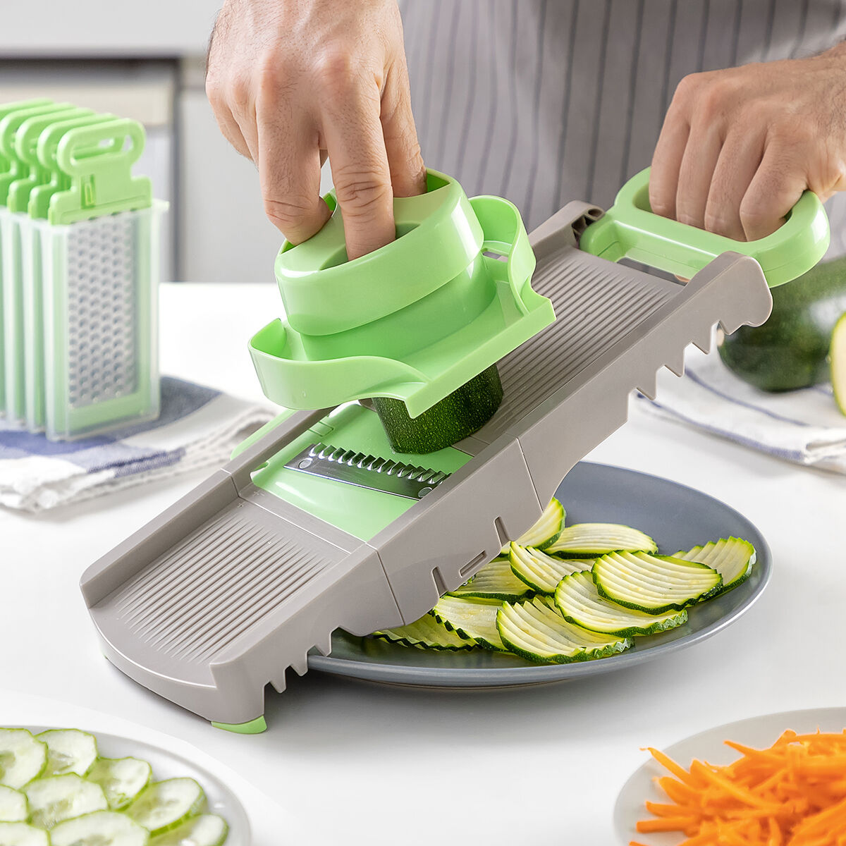 Una persona affetta zucchine con una mandolina verde e grigia. Zucchine a fette su un piatto. Si vedono altre verdure e una grattugia.