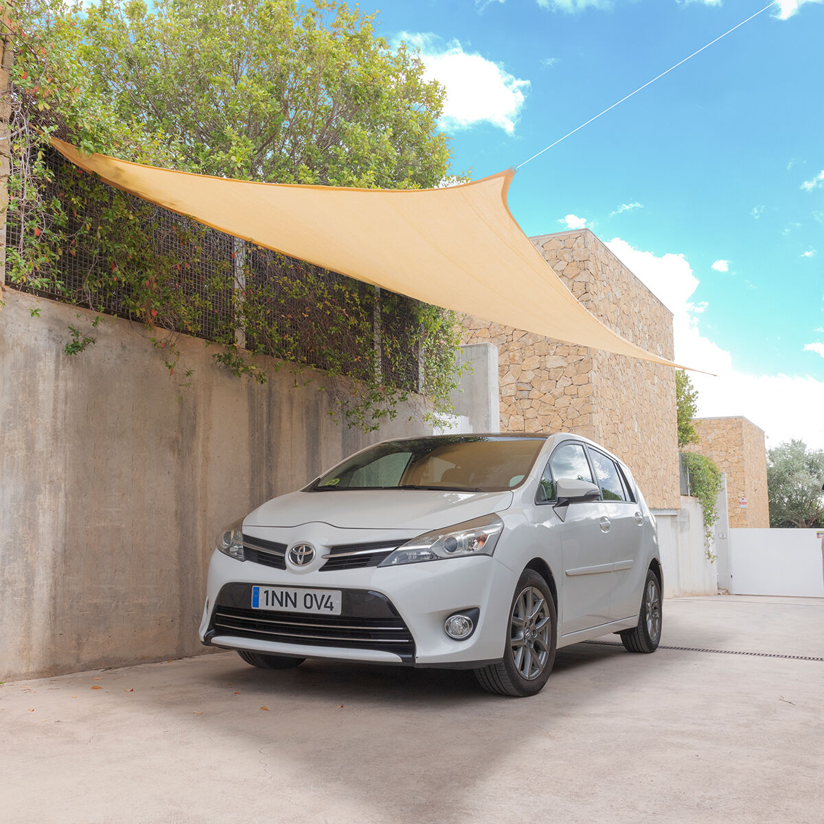 Weißer Toyota unter einem beigen Sonnensegel geparkt. Steinmauer und Laub im Hintergrund, blauer Himmel.