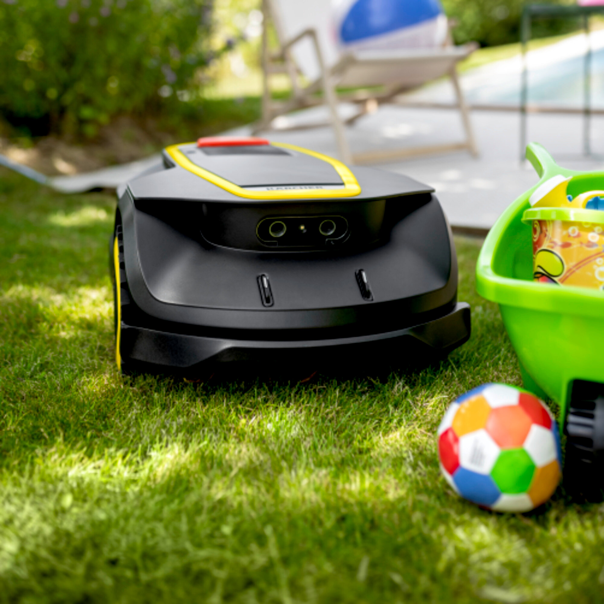 Czarny robot koszący na zielonej trawie, w pobliżu zielonego wózka zabawkarskiego i kolorowej piłki, z rozmytym tłem.