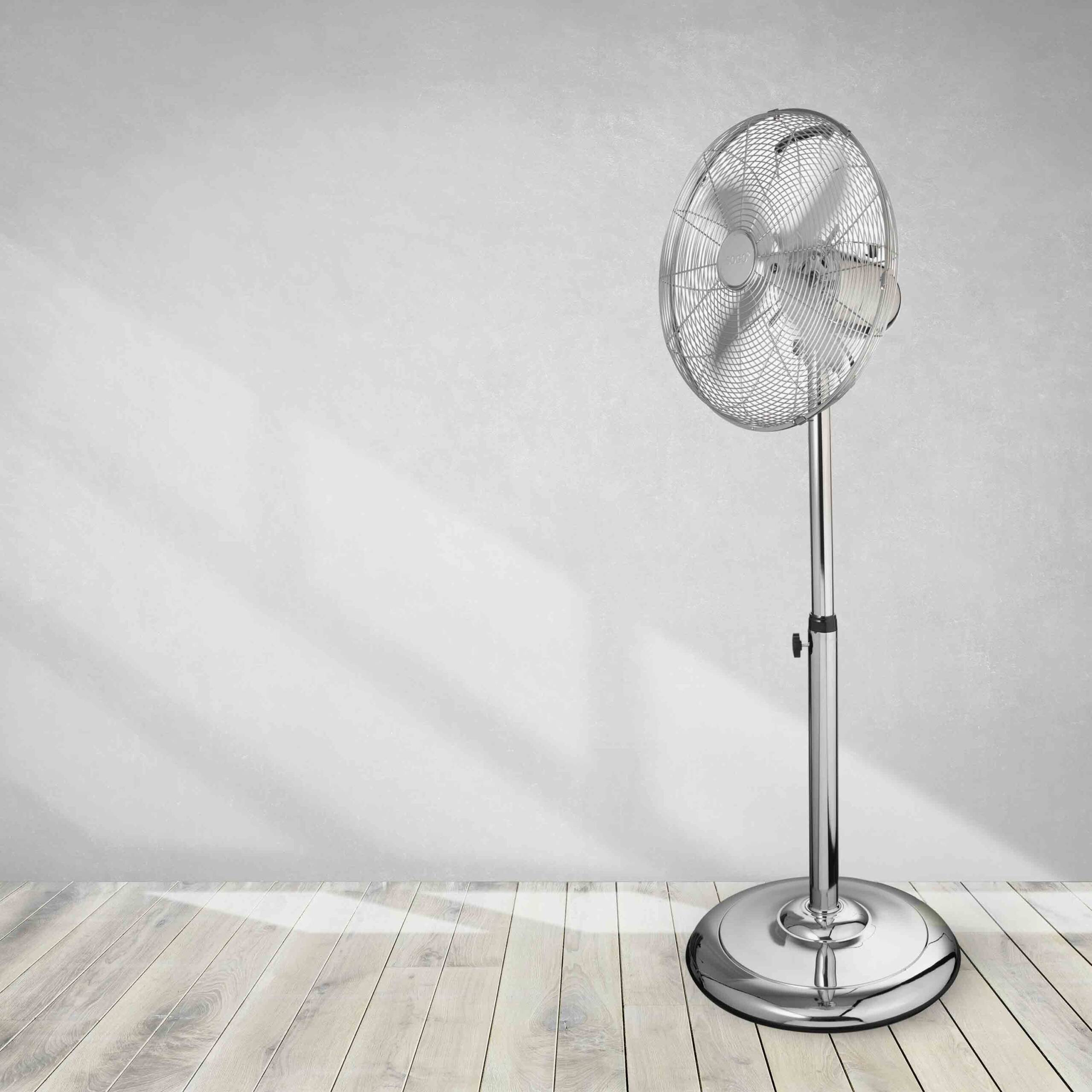 Un ventilador de pedestal cromado está sobre un piso de madera contra una pared gris clara. El ventilador está encendido.