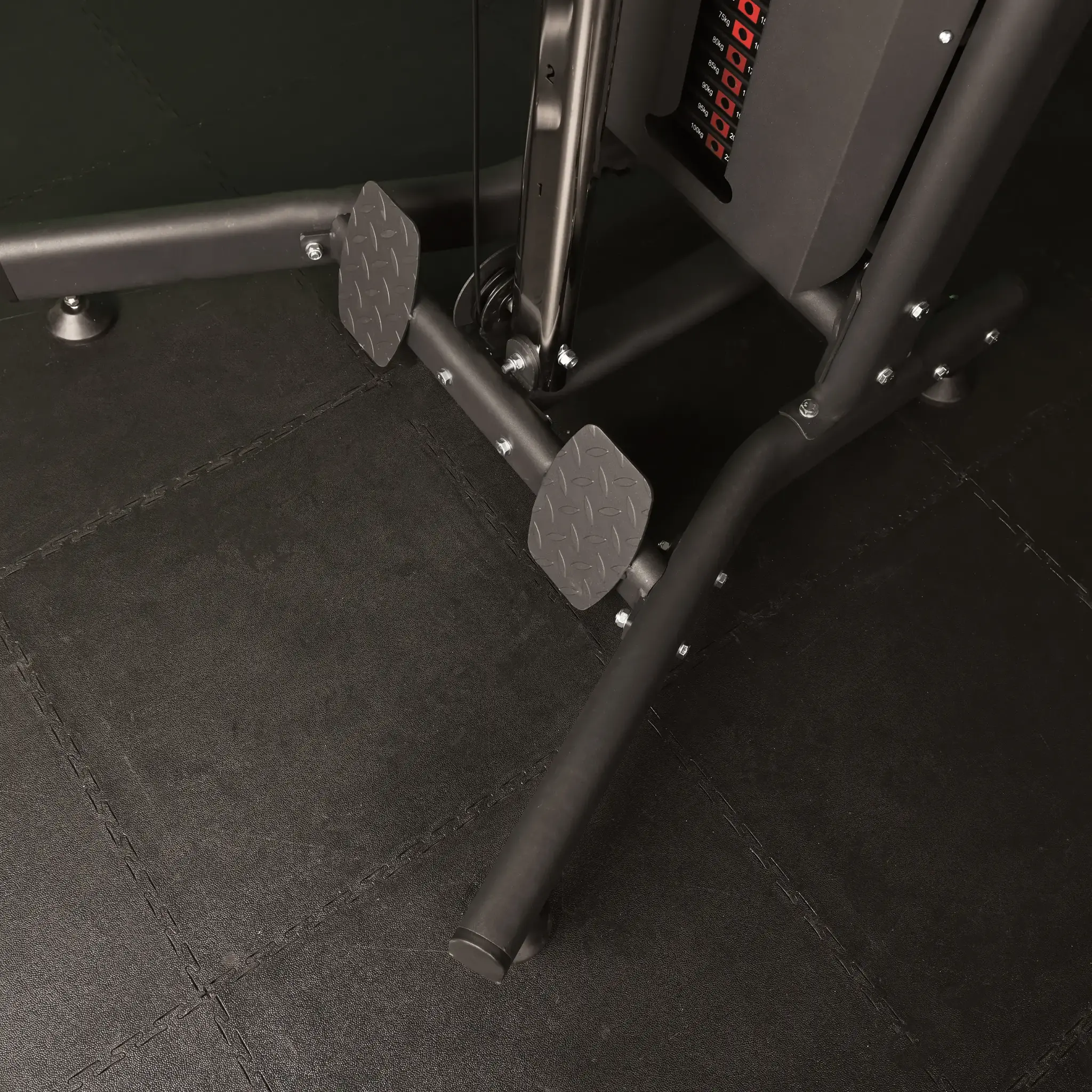 A gym weight machine's base, with textured foot platforms. The floor is black, with weight indicators on the side.