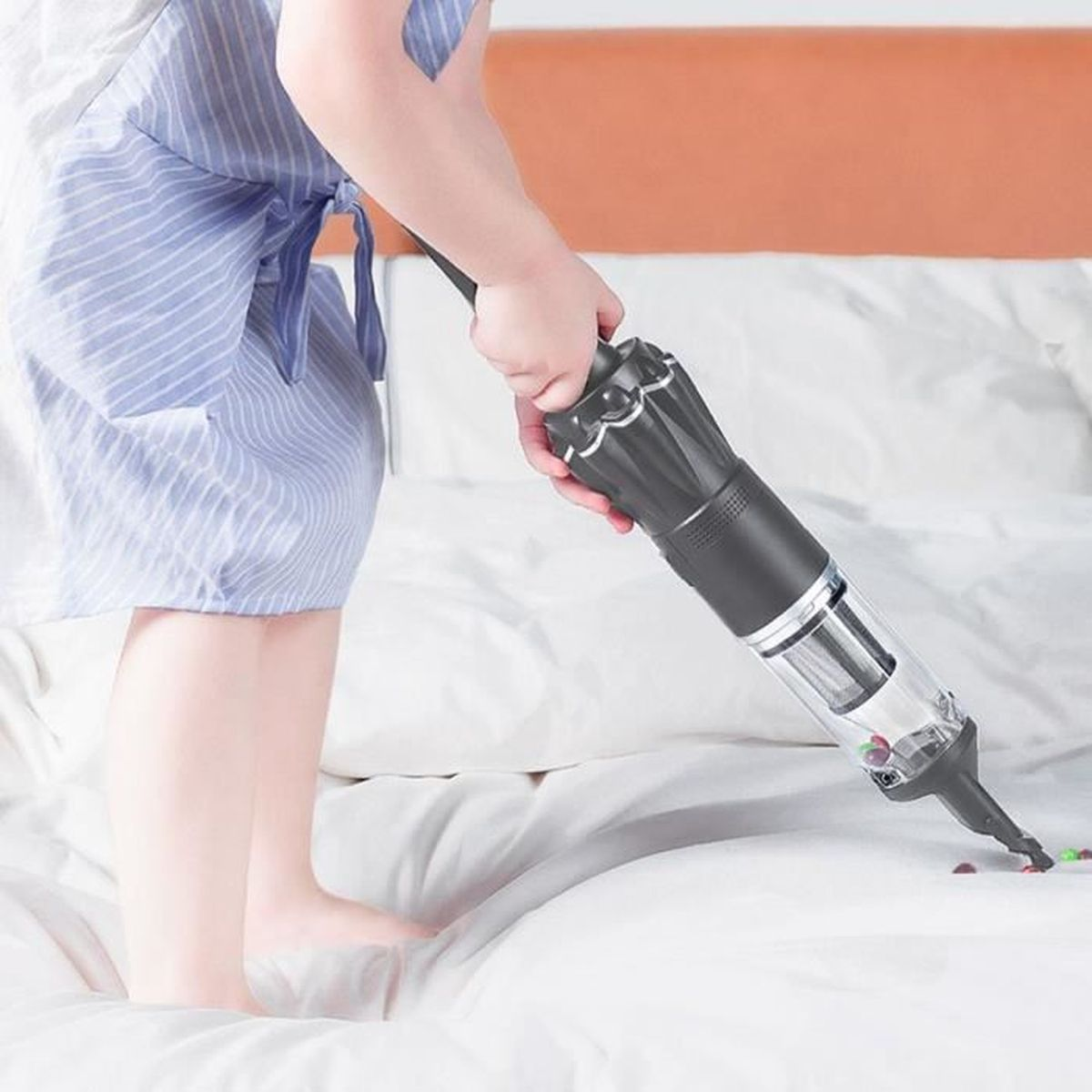 Una persona in abito blu sta passando l'aspirapolvere su un letto bianco con un aspirapolvere grigio. Lo sfondo mostra una testiera marrone chiaro.