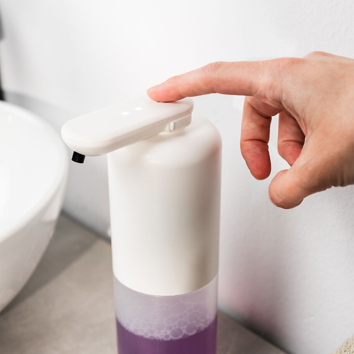 Eine Hand drückt auf einen weißen Seifenspender. Der Spender steht auf einer grauen Oberfläche, im Hintergrund ein Waschbecken. Lila Flüssigkeit sichtbar.