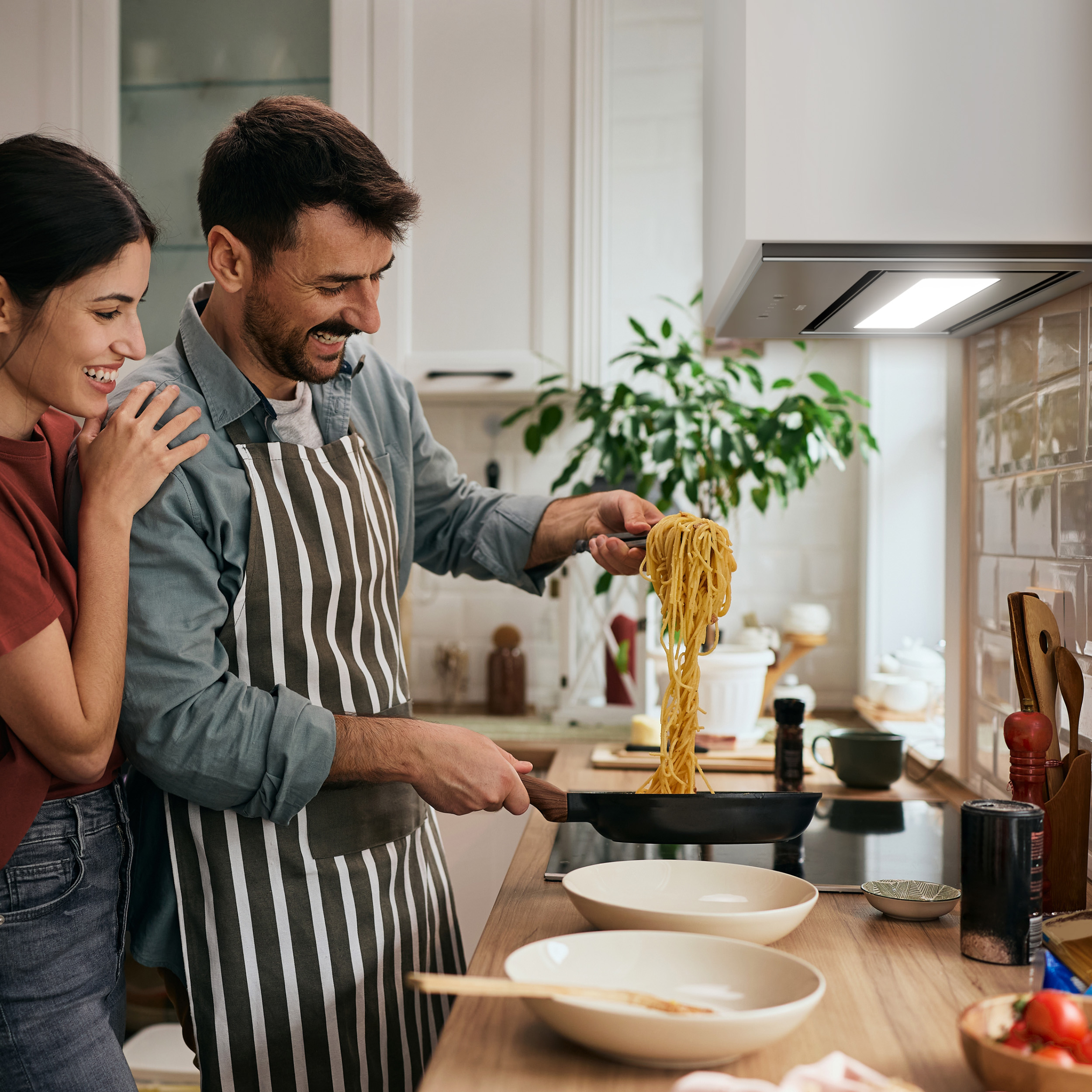 Een stel in een keuken, man houdt pasta uit een pan. Vrouw glimlacht met hand op zijn schouder. Keuken achtergrond.