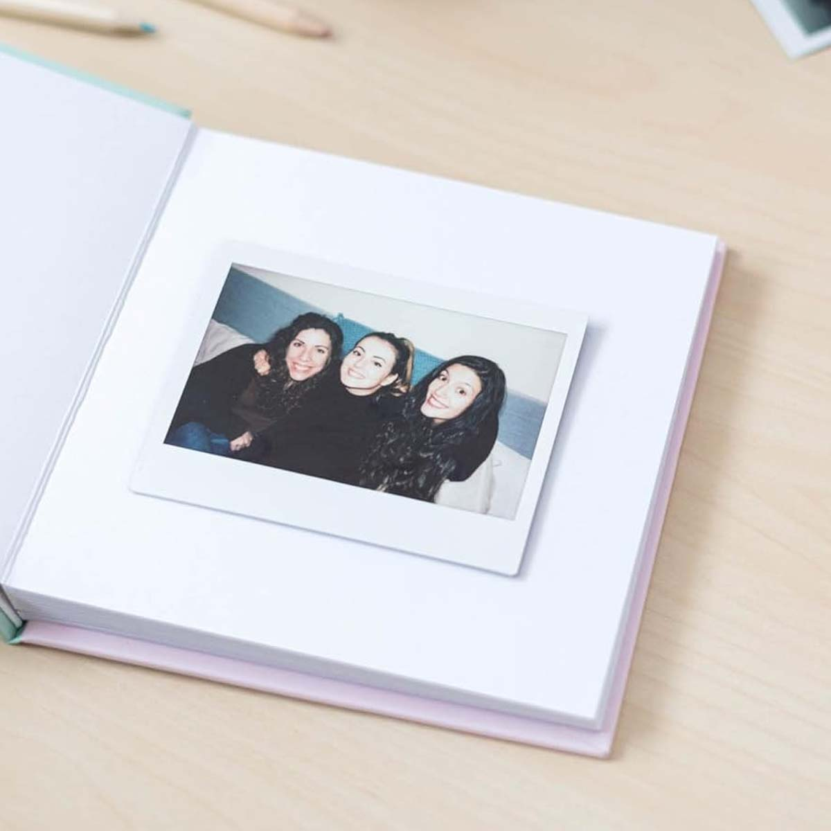 Un album photo avec une photo Polaroid de trois femmes souriantes. L'album est ouvert sur une table en bois avec des crayons.