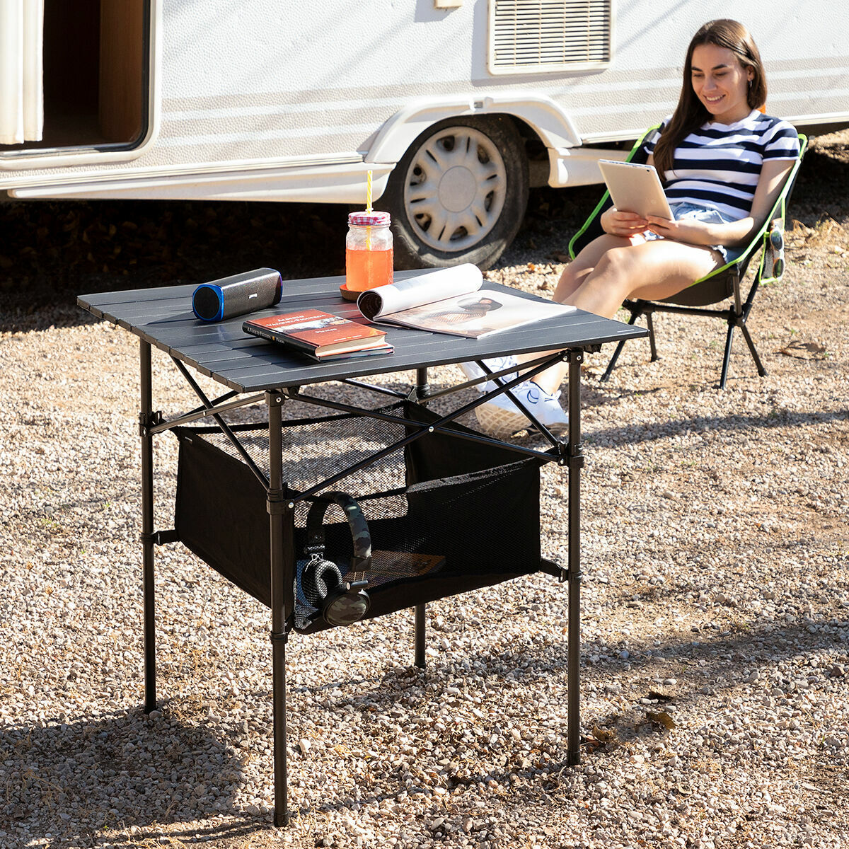 Frau benutzt Tablet neben einem Campingtisch mit Getränken, Büchern und einem Lautsprecher. Im Hintergrund ein Wohnwagen.
