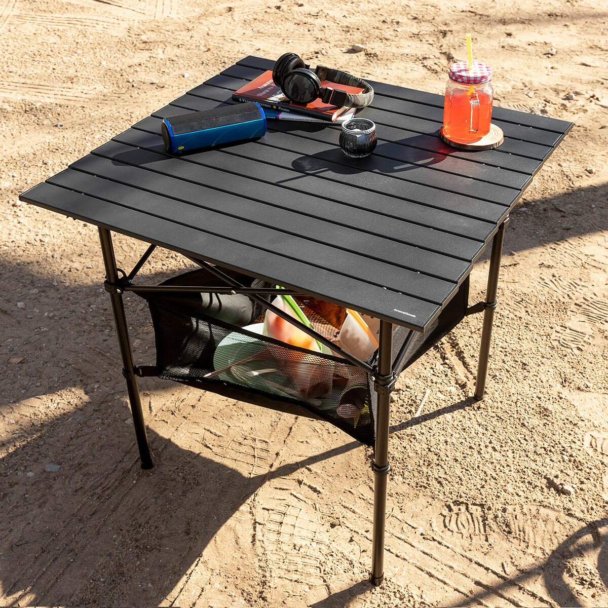 Ein schwarzer Campingtisch auf Sand trägt Kopfhörer, einen Lautsprecher, ein Getränk und andere Gegenstände. Darunter befindet sich ein Netzregal.