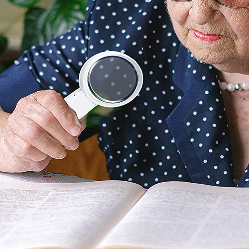 Eine ältere Person benutzt eine Lupe, um ein Buch zu lesen. Sie trägt eine Brille und ein Polka-Dot-Hemd und konzentriert sich auf den Text.